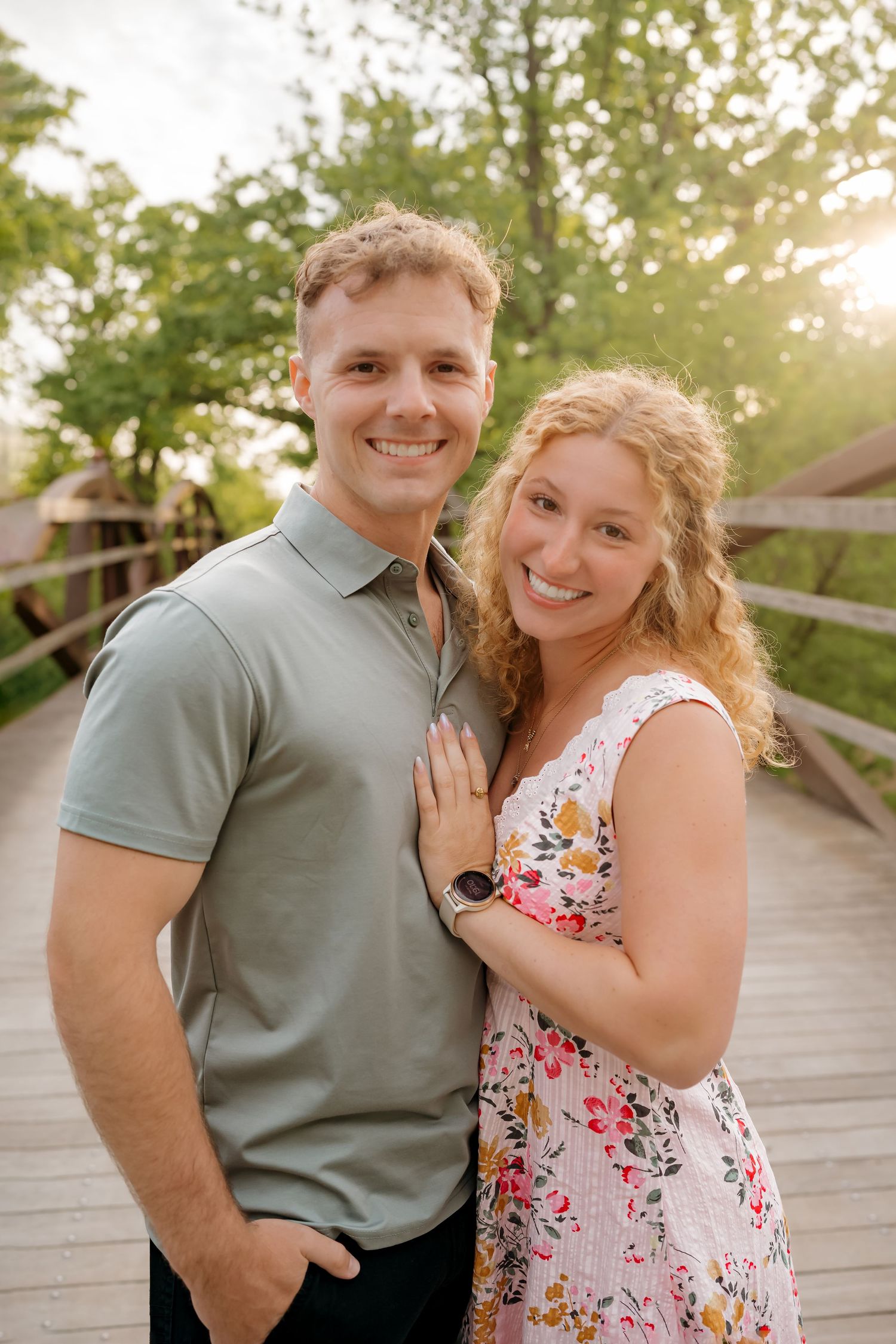 A smiling couple poses on a wooden bridge surrounded by green trees in warm golden sunlight.