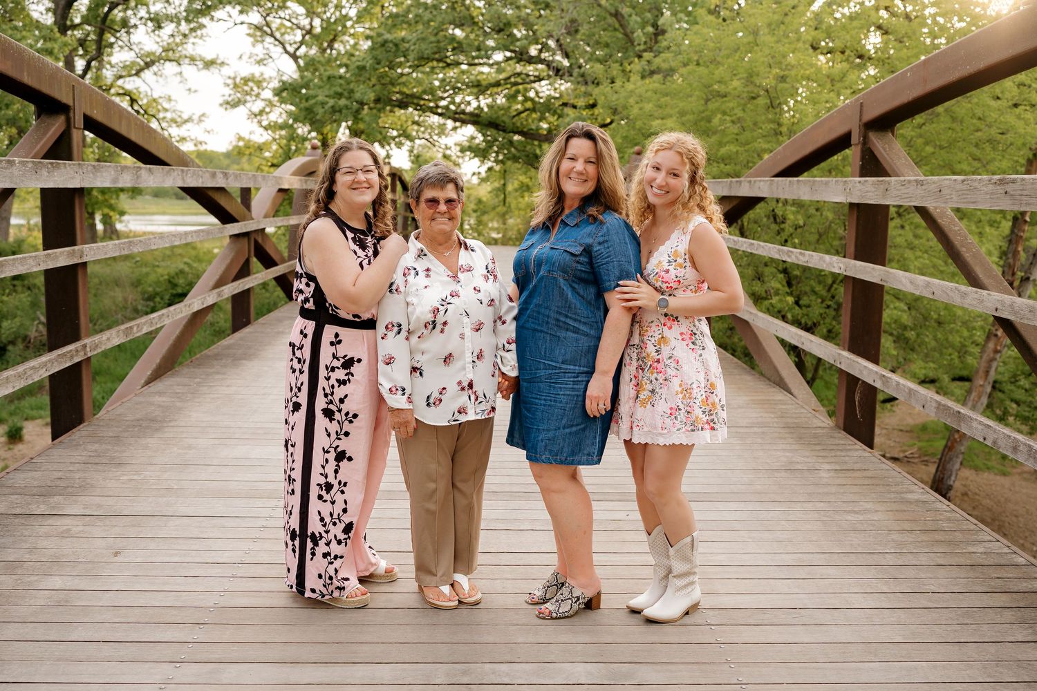 Four women smiling together on a wooden bridge surrounded by lush green trees in an outdoor portrait.