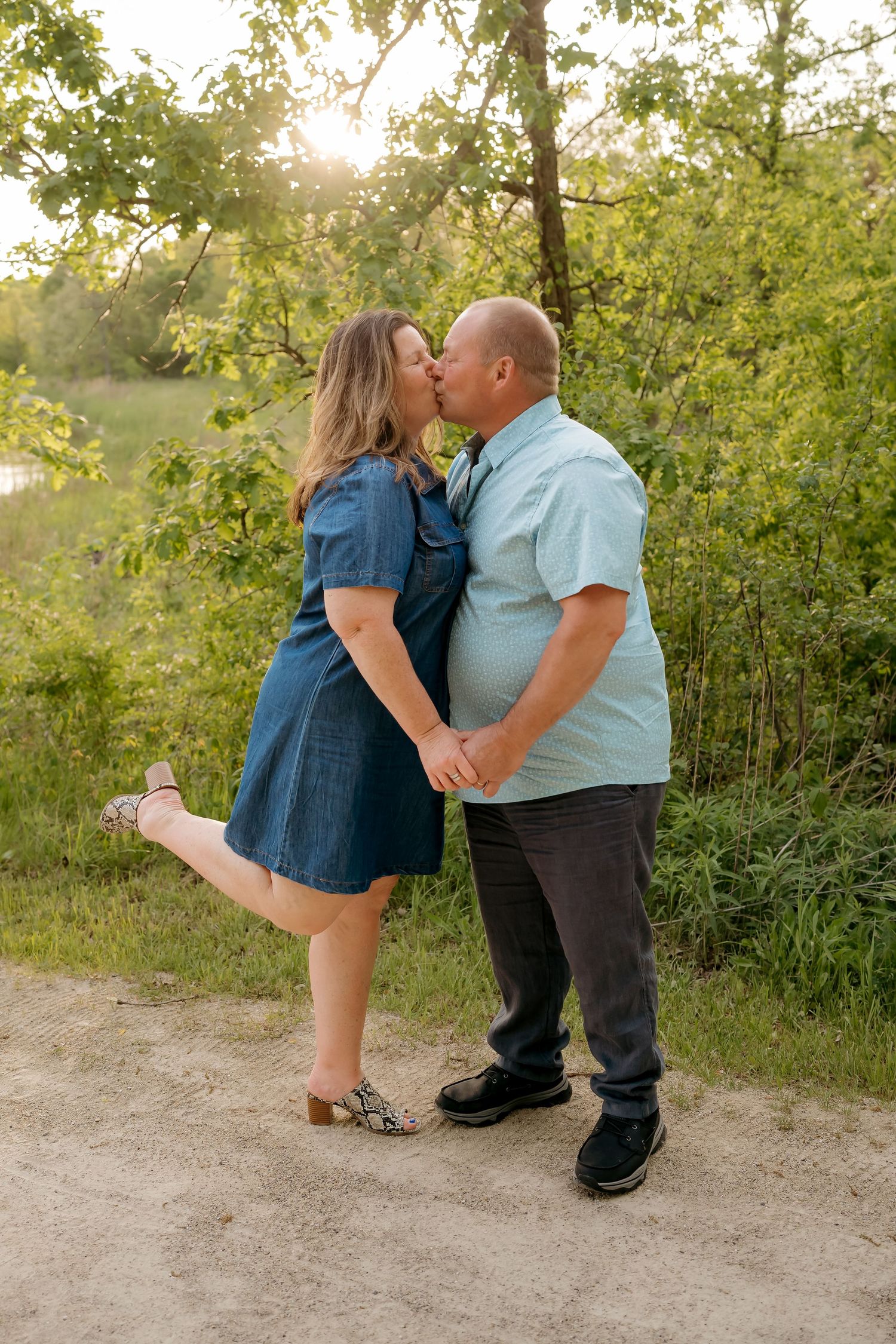 Couple sharing a romantic kiss outdoors on a dirt path, surrounded by lush green trees in golden hour light.