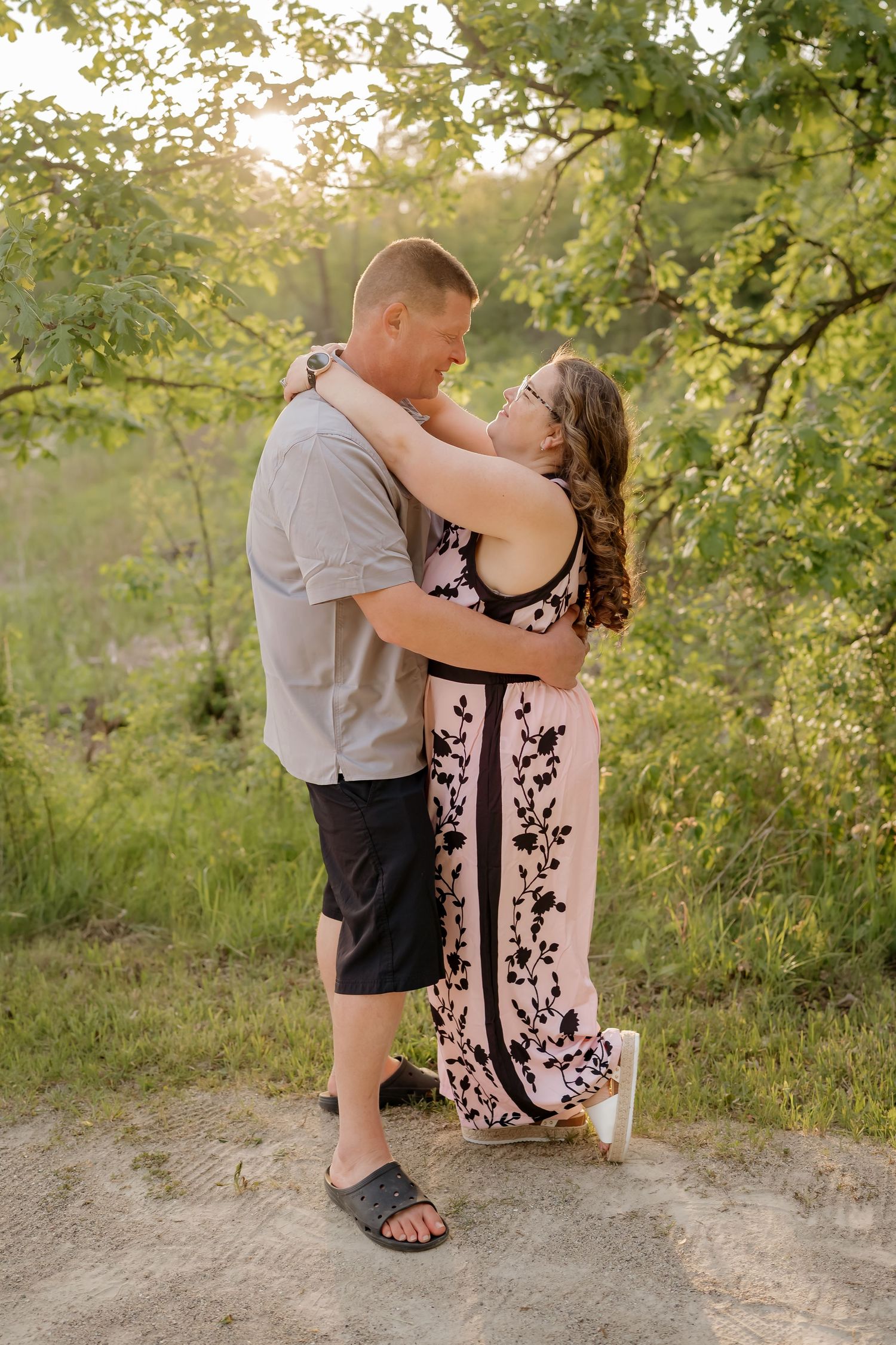 Couple embracing outdoors at golden hour, woman in floral dress, man in casual shorts, surrounded by lush green trees.