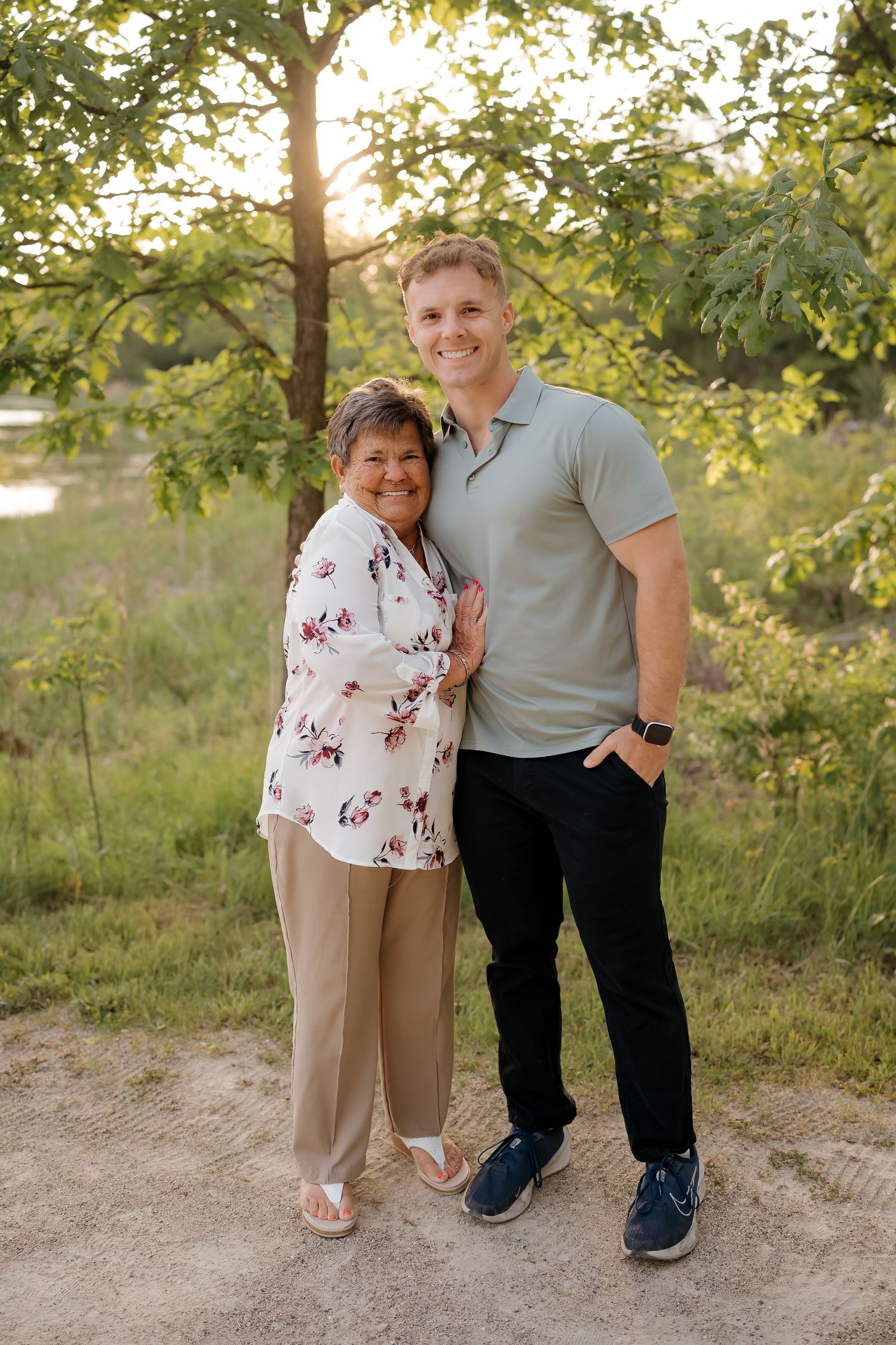 A young man and older woman smile together outdoors under green trees in warm golden hour light.
