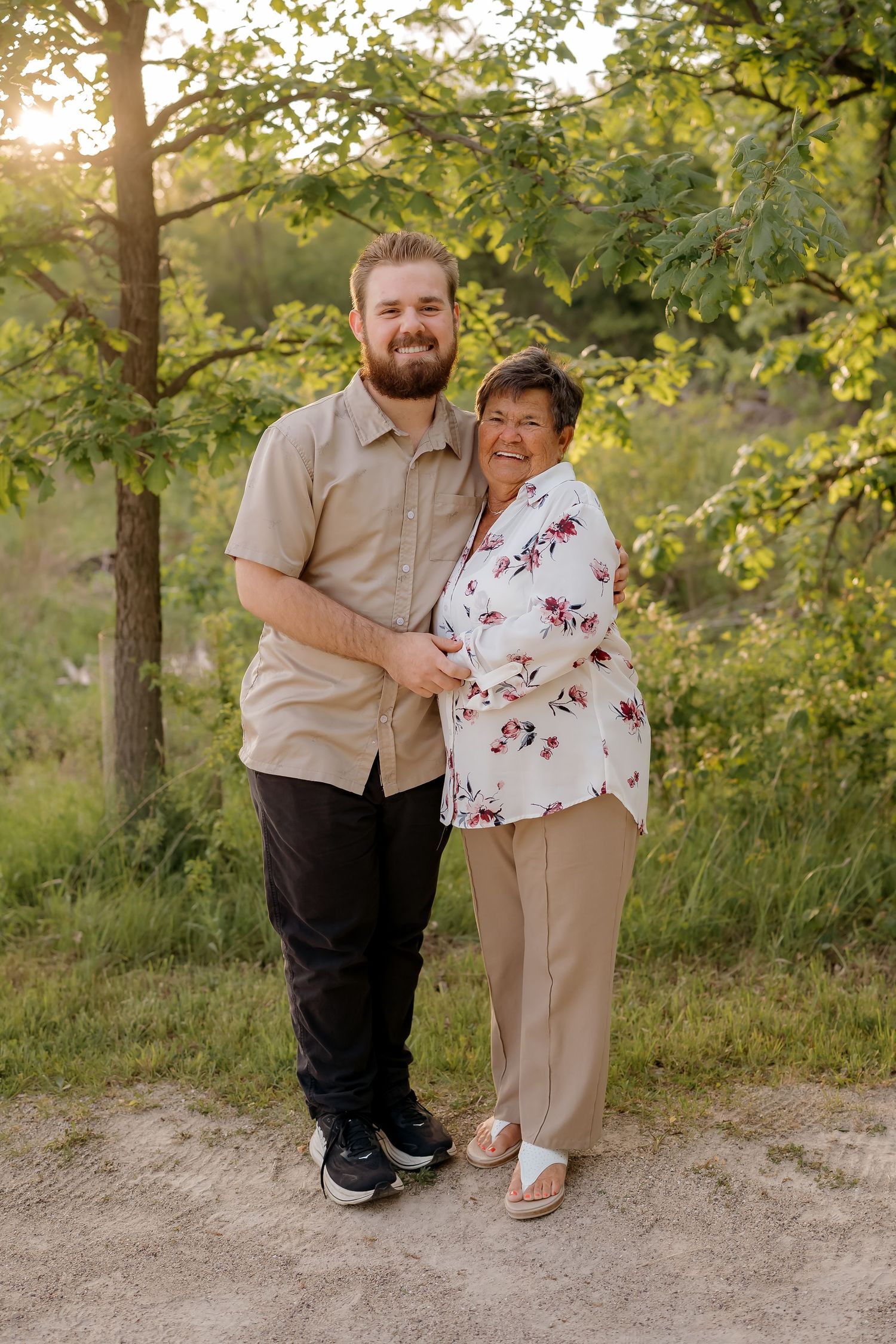 A young man and older woman smile together outdoors at golden hour, surrounded by lush green trees.