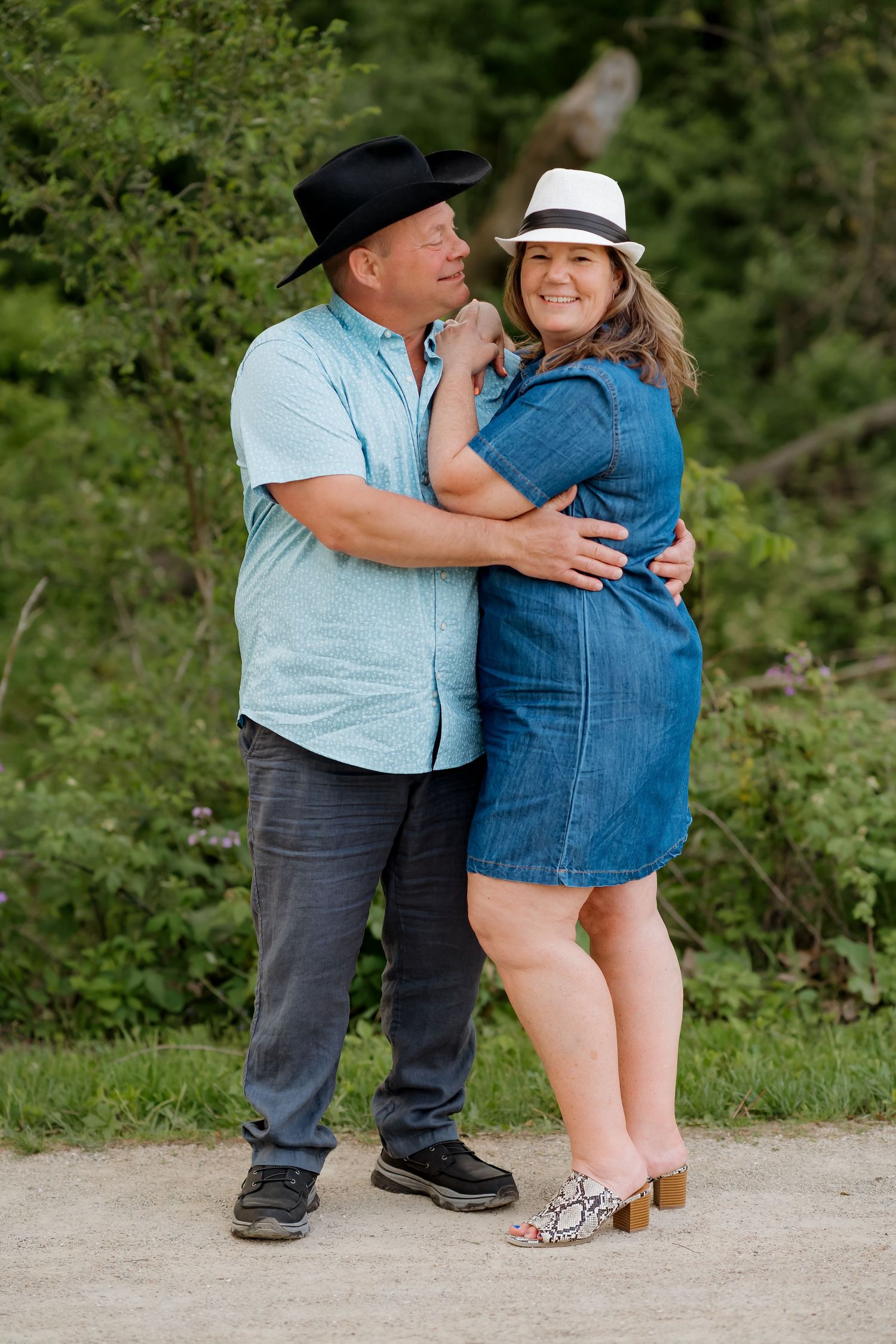Couple embracing outdoors, man in black cowboy hat and blue shirt, woman in denim dress and white fedora.