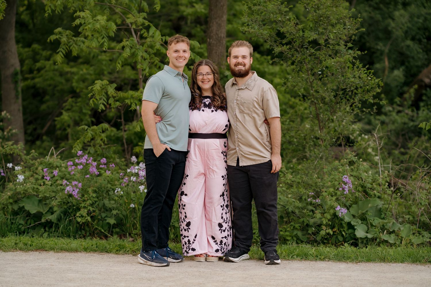 Three people smiling and posing together outdoors on a path surrounded by lush green trees and purple flowers.