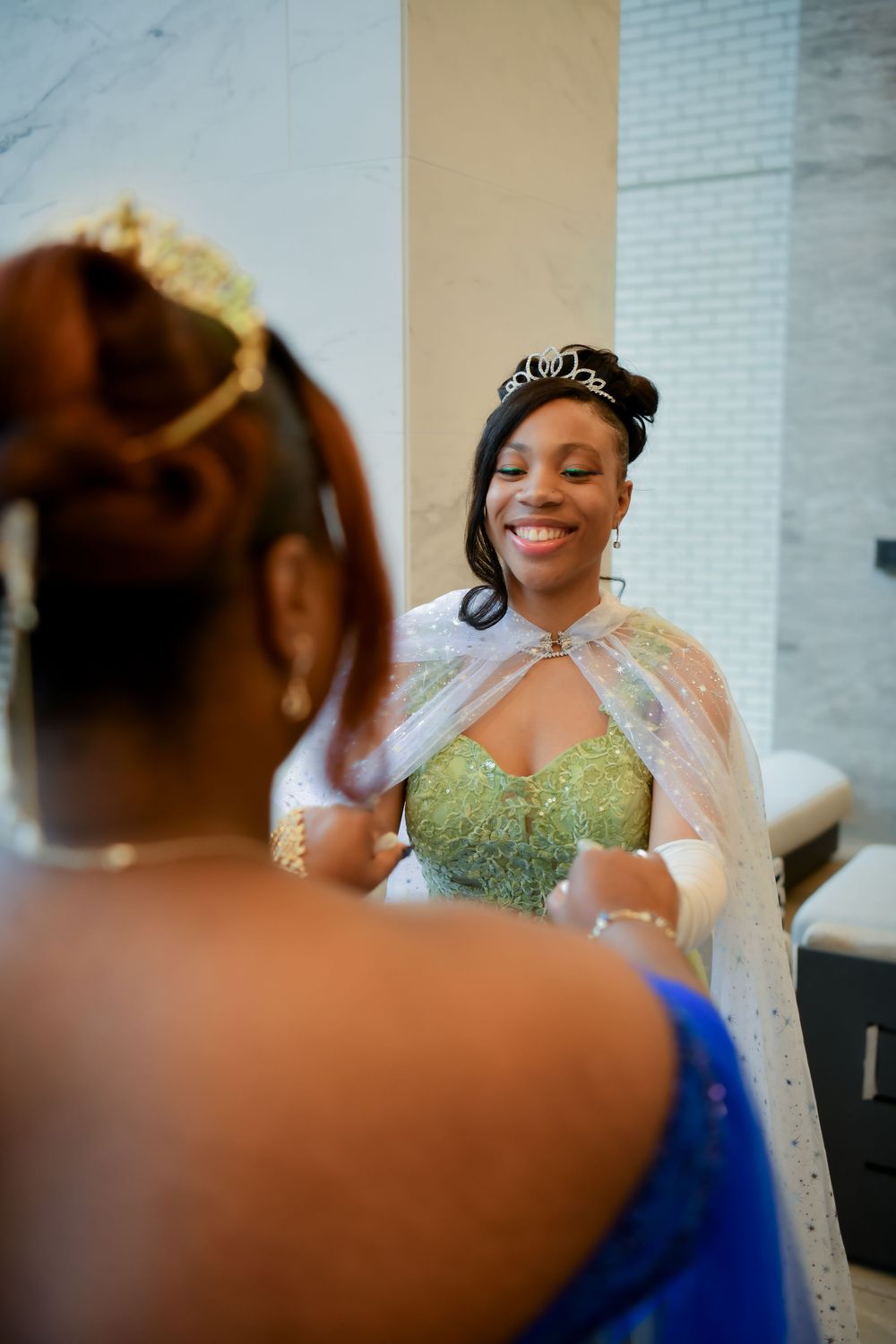 Young woman wearing tiara and green sequin gown with white cape smiles in mirror reflection indoors.