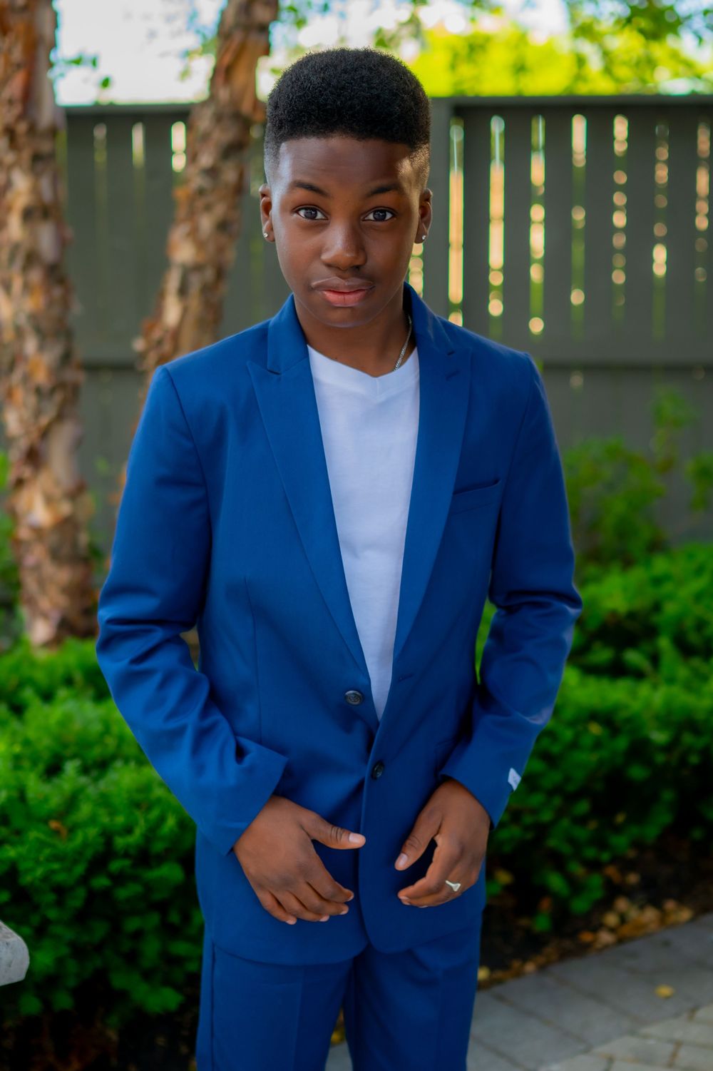 Young man in a blue suit and white shirt posing outdoors with green shrubs and wooden fence backdrop.