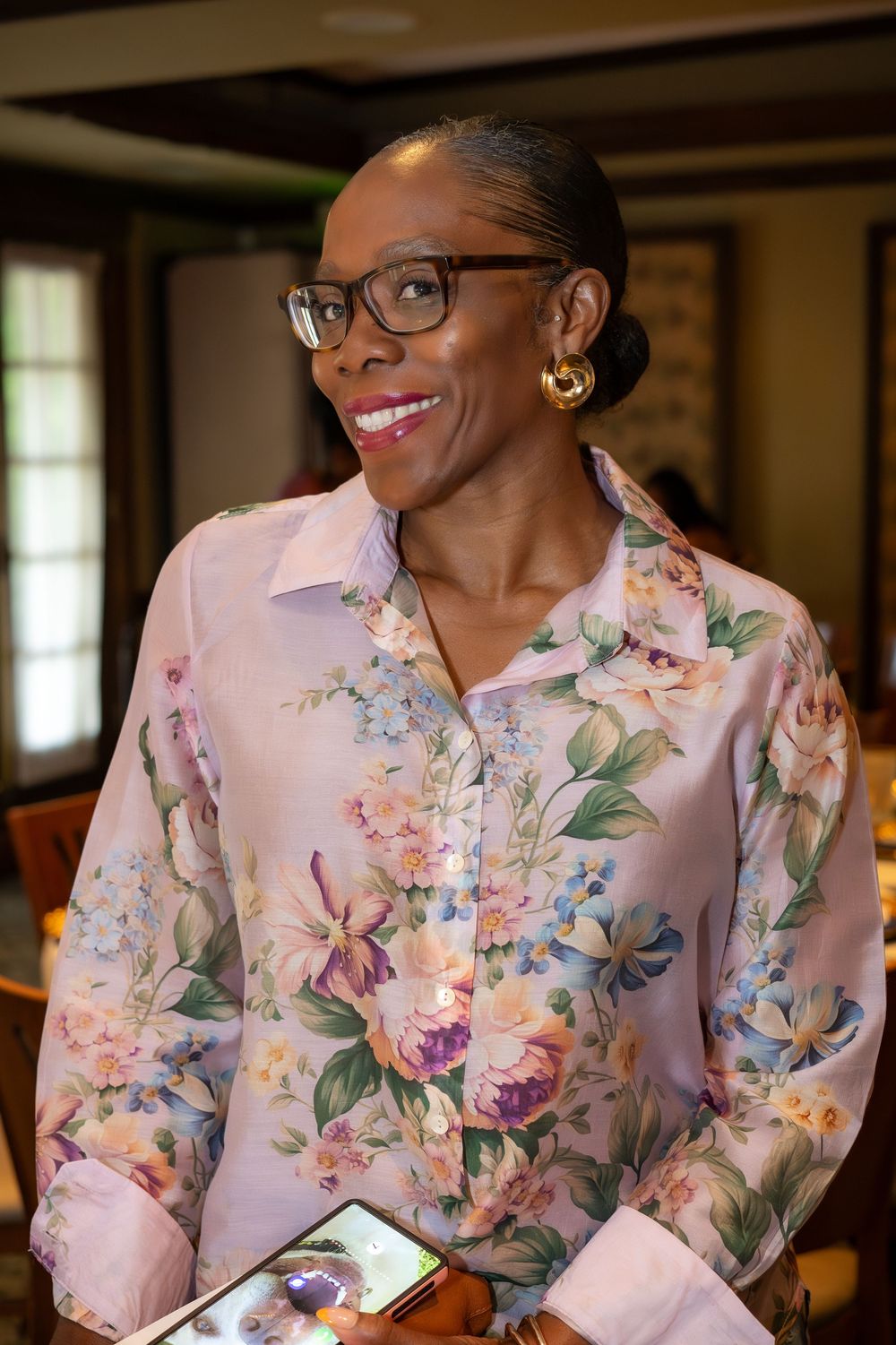 Elegant woman in floral blouse wearing glasses smiles warmly while holding a phone at an indoor event.