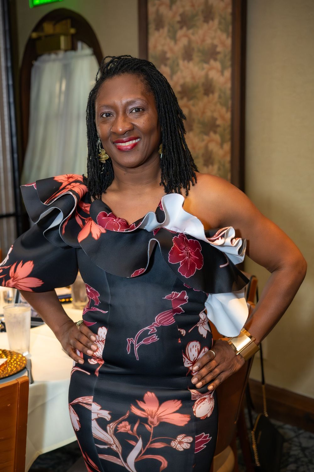 Woman in black floral off-shoulder ruffled dress poses confidently with hands on hips at indoor event.