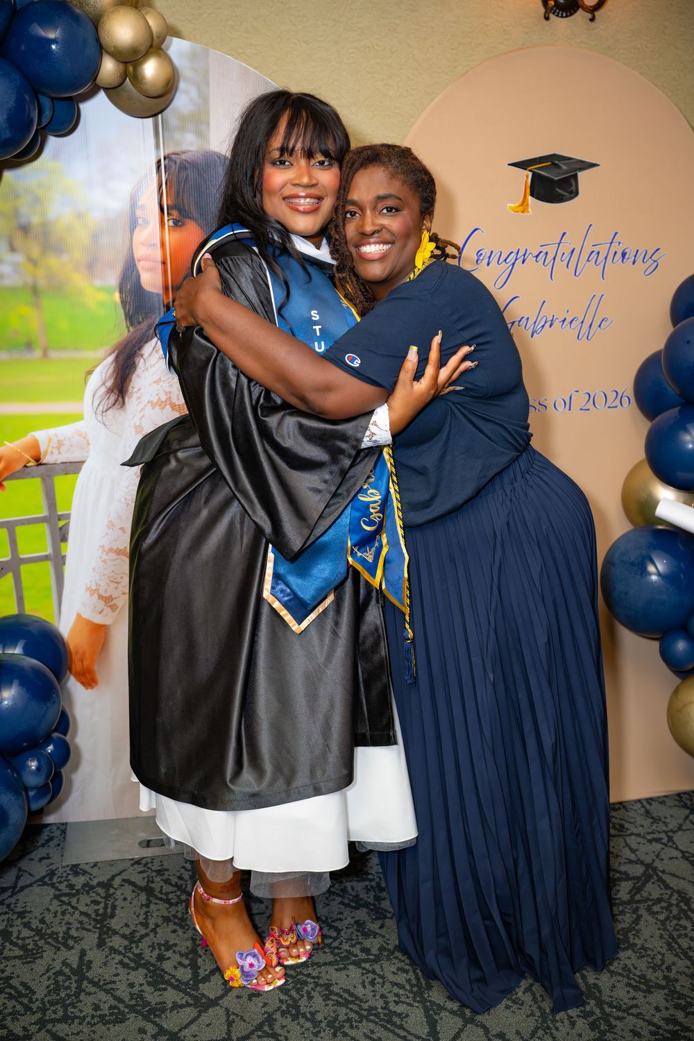 Graduate in cap and gown shares a warm hug with a woman in blue dress at a Congratulations Gabrielle party.