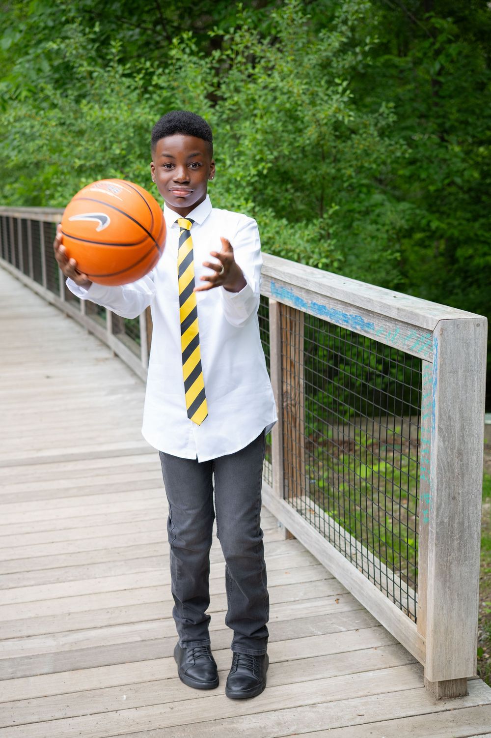 Young boy in white shirt and striped tie extends basketball toward camera on a wooden bridge outdoors.