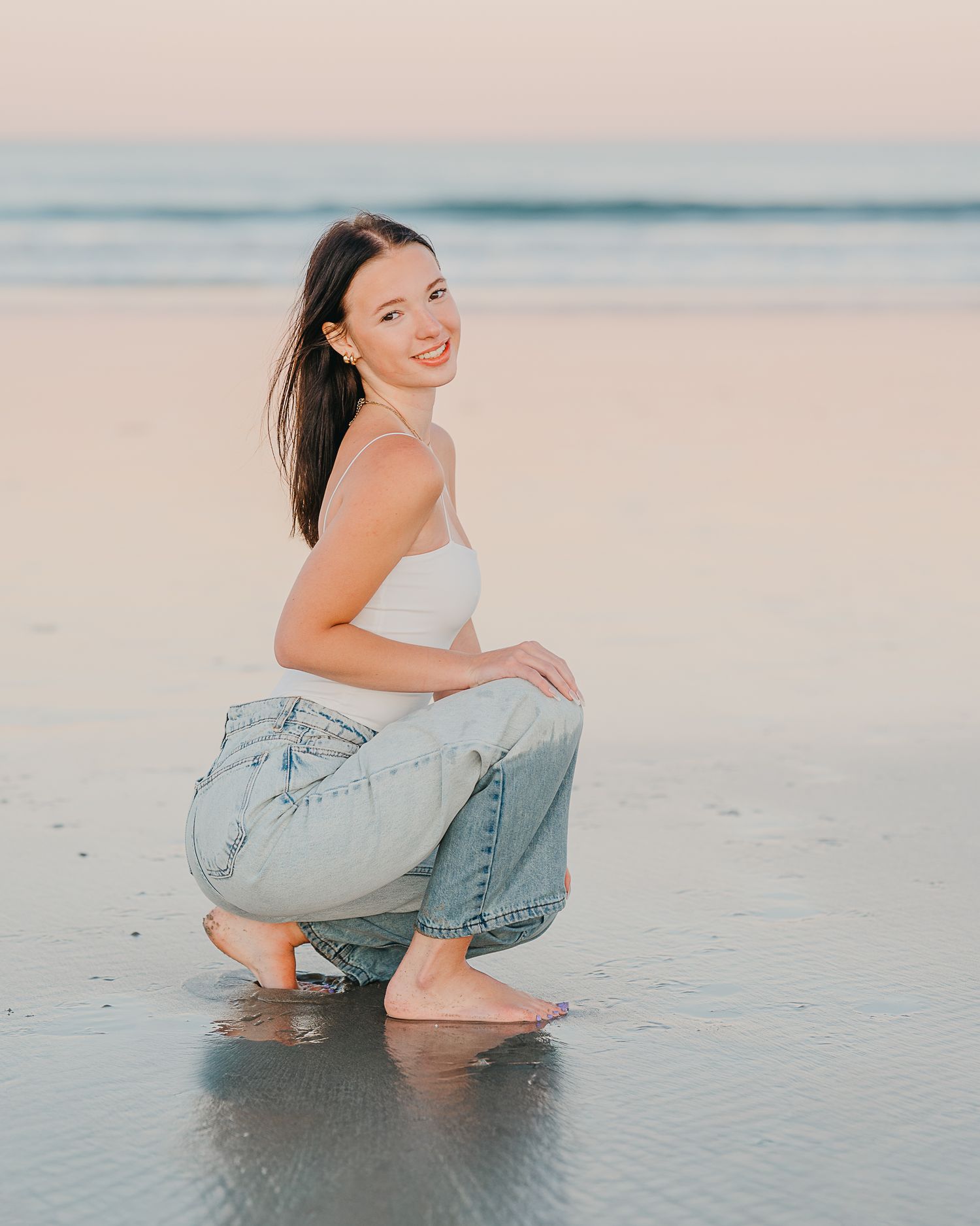 high school senior in white crop top and jeans crouching on a sandy beach at sunset, smiling over her shoulder.