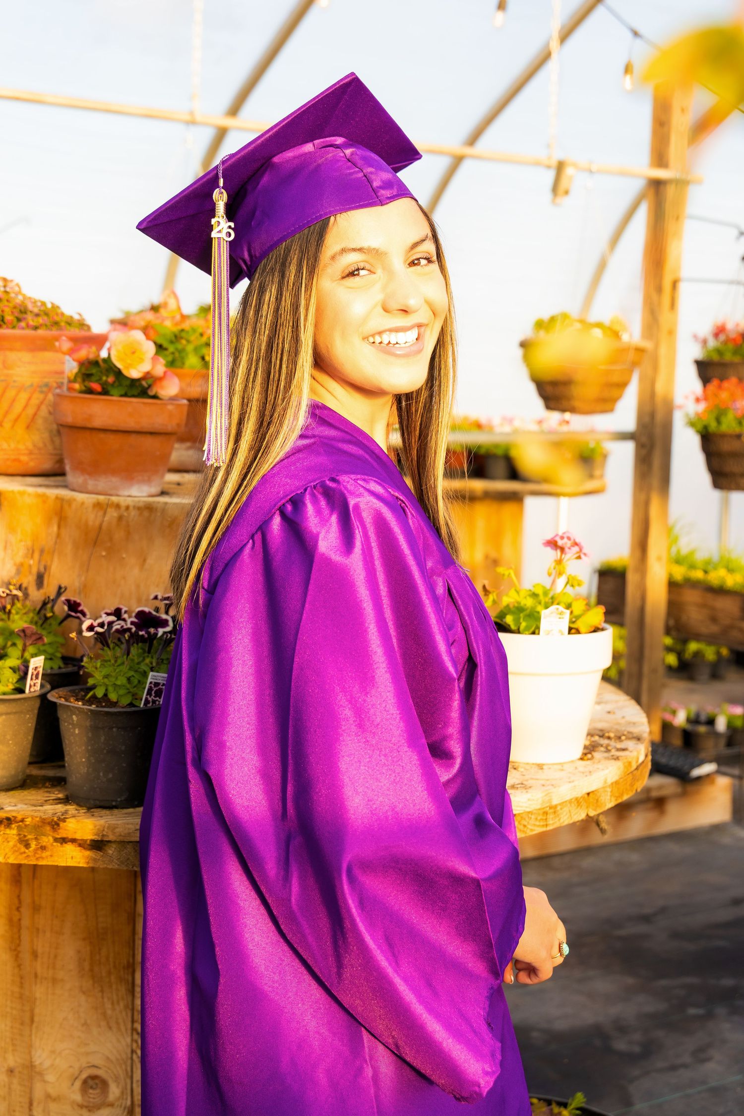 Smiling young woman in purple graduation cap and gown posing in a greenhouse surrounded by colorful potted plants in Connell,Wa.