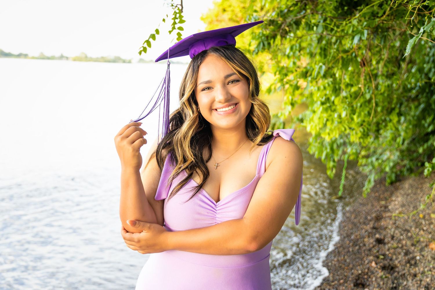 Young woman in lavender dress wearing purple graduation cap smiles outdoors near water with lush green trees in Kennewick, Wa.