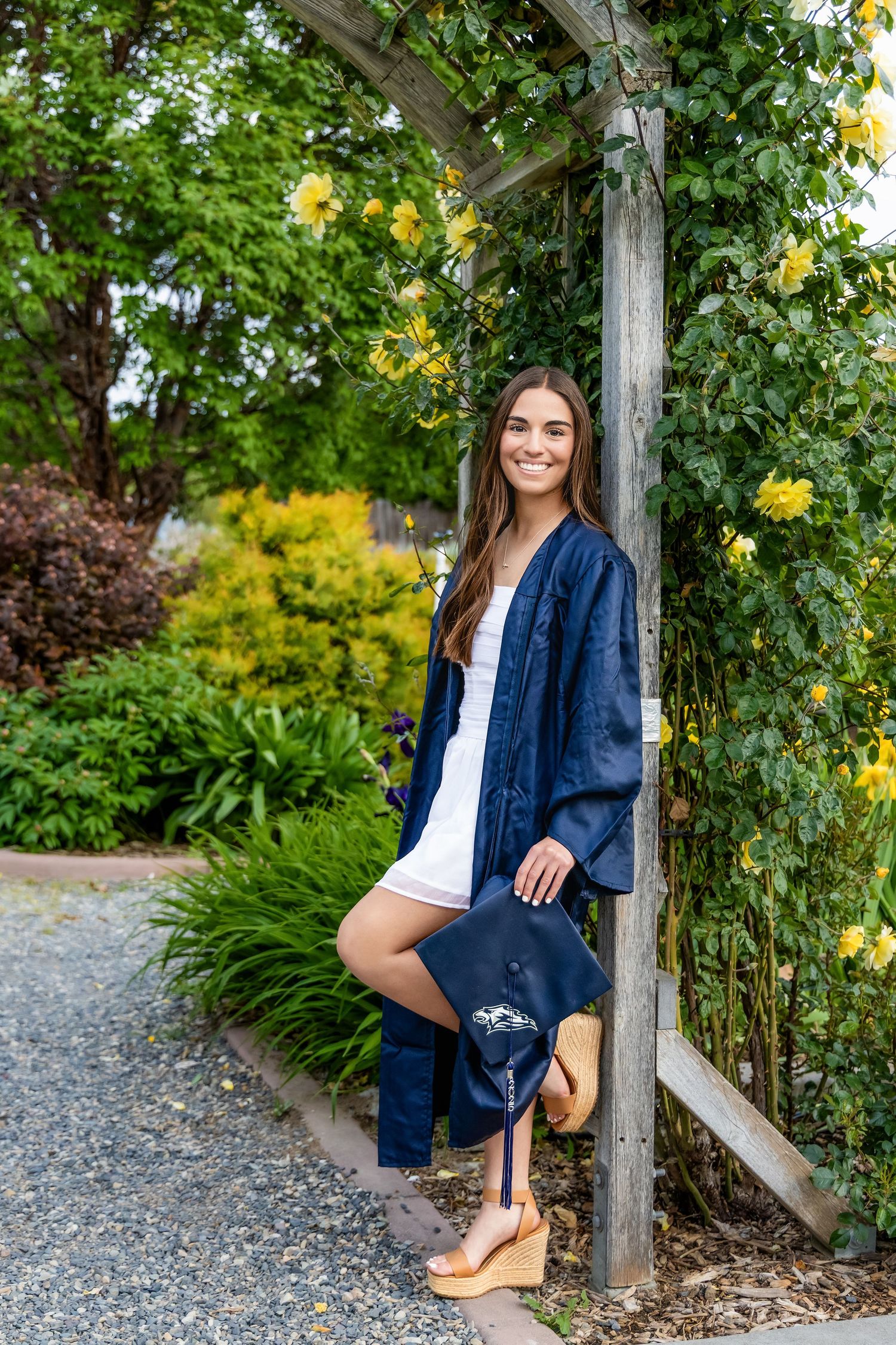 Young woman in navy graduation gown and cap smiling under a wooden garden arbor with yellow flowers in Kennewick, Wa.
