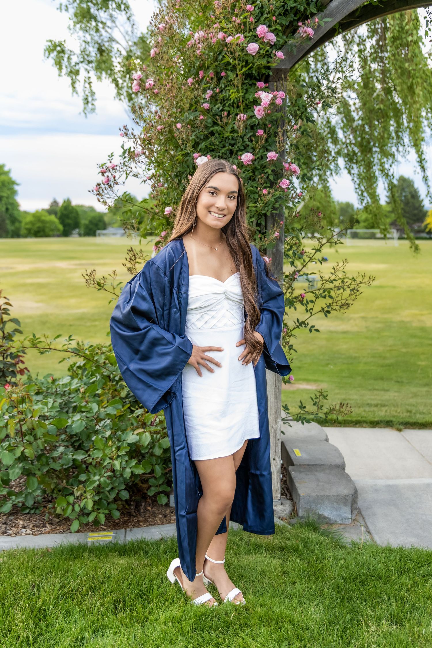 Smiling graduate in navy blue cap gown and white dress poses outdoors near flowering arch in green park in Kennewick, Wa.