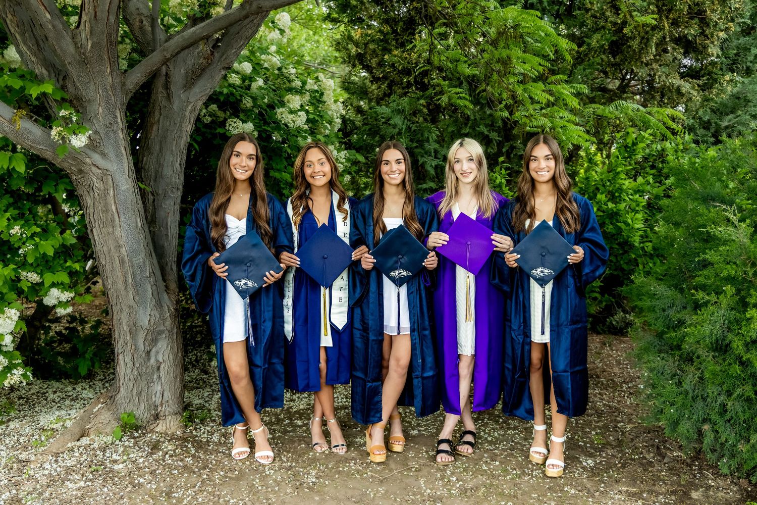 Five female graduates in navy blue caps and gowns pose outdoors under a tree holding diplomas in Kennewick, Wa.