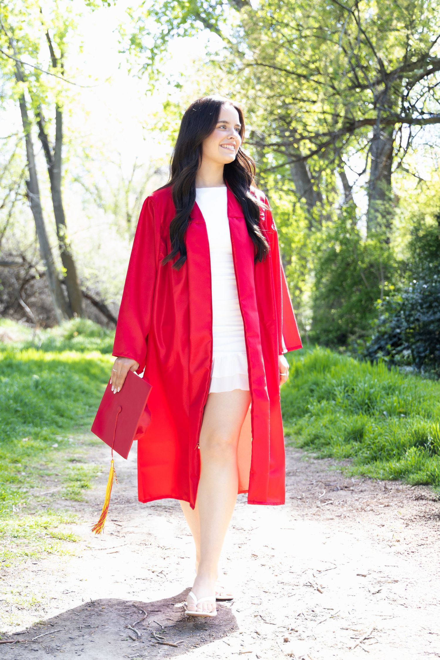 Smiling young woman in red graduation cap and gown walking outdoors on a sunny tree-lined path in Kennewick, Wa.