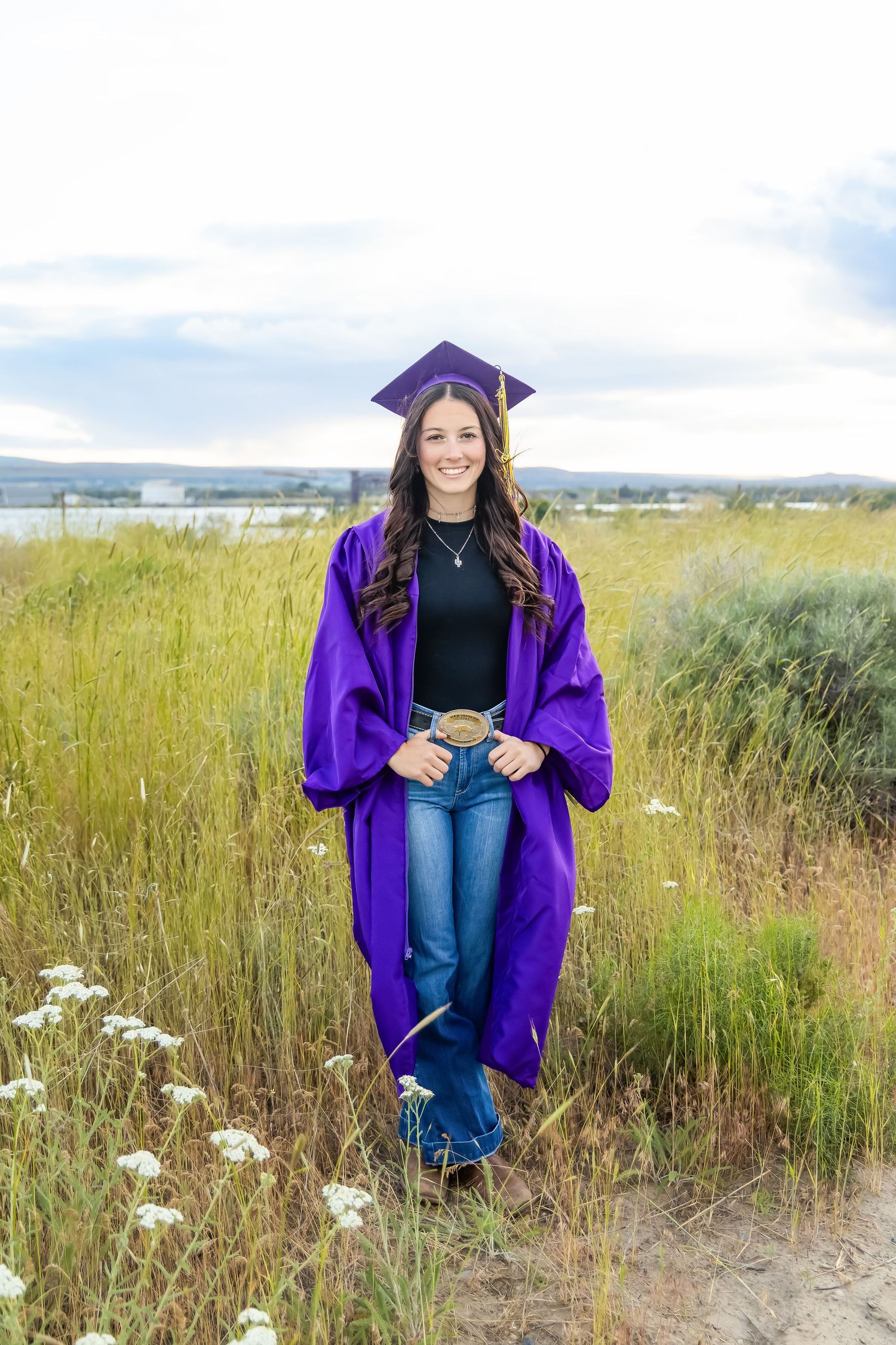 Young woman in purple graduation cap and gown smiling outdoors in a field, holding a medallion, wearing jeans in Burbank, Wa.