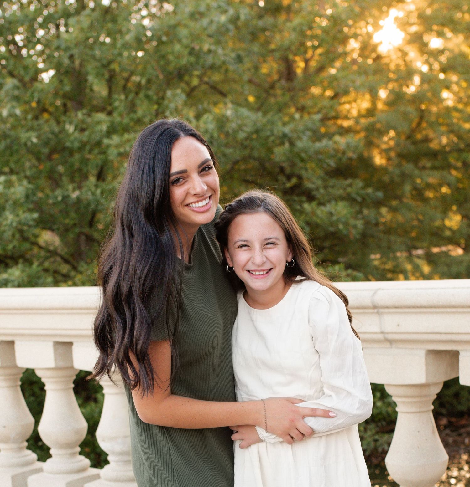 Two smiling females, older girl in green dress hugging younger girl in white dress, posing outdoors near stone balustrade.
