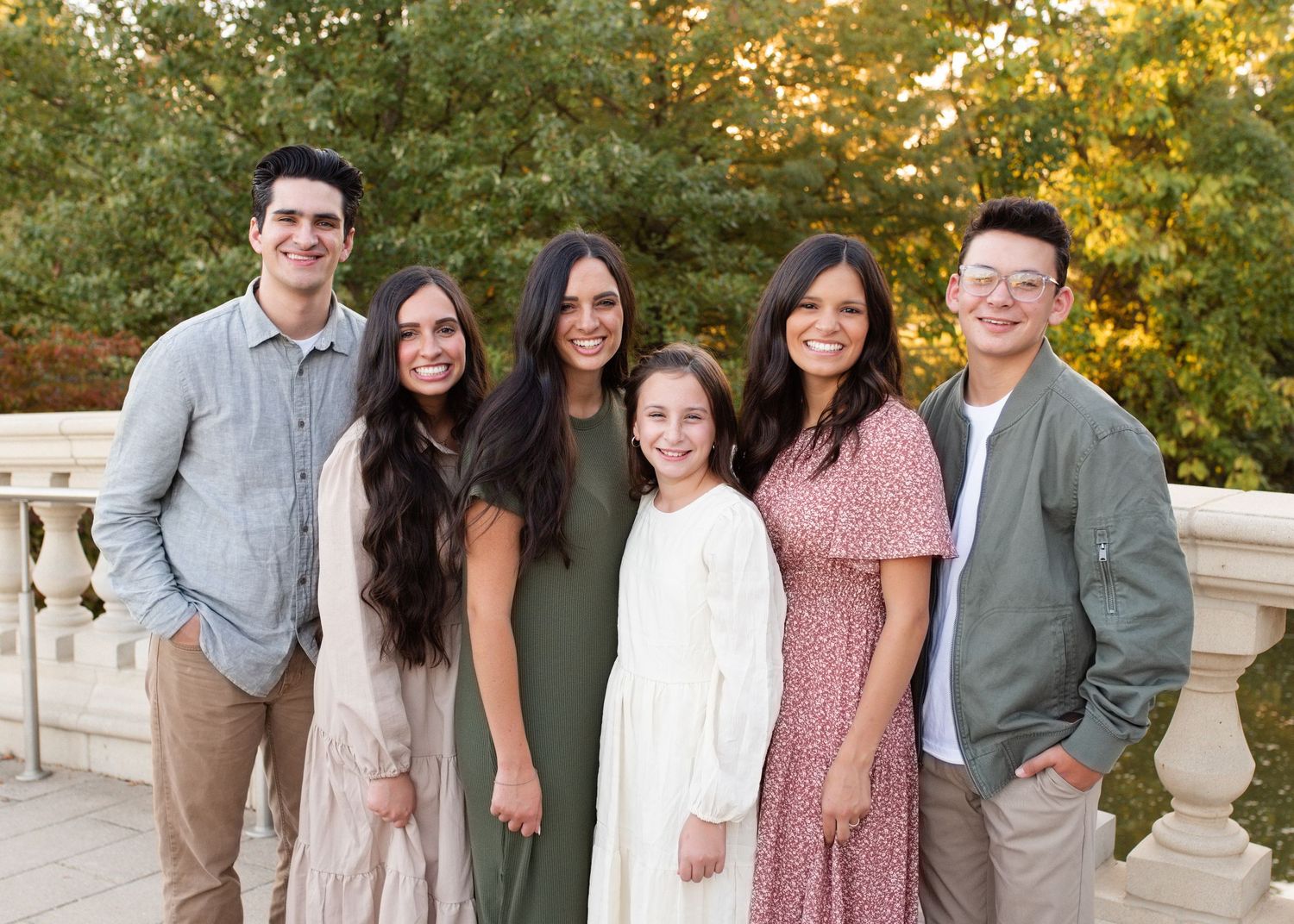 Six young adults smiling together outdoors on a stone terrace surrounded by lush green trees.