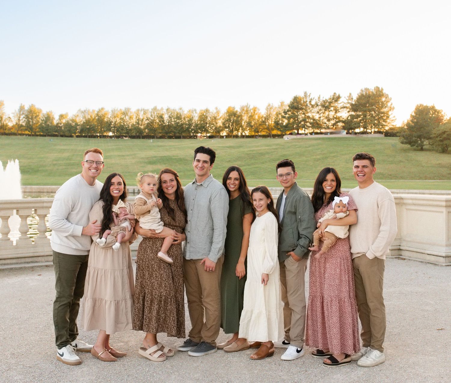 Large family group portrait outdoors at golden hour, smiling adults posing on stone terrace with lush green landscape at forest park in STL