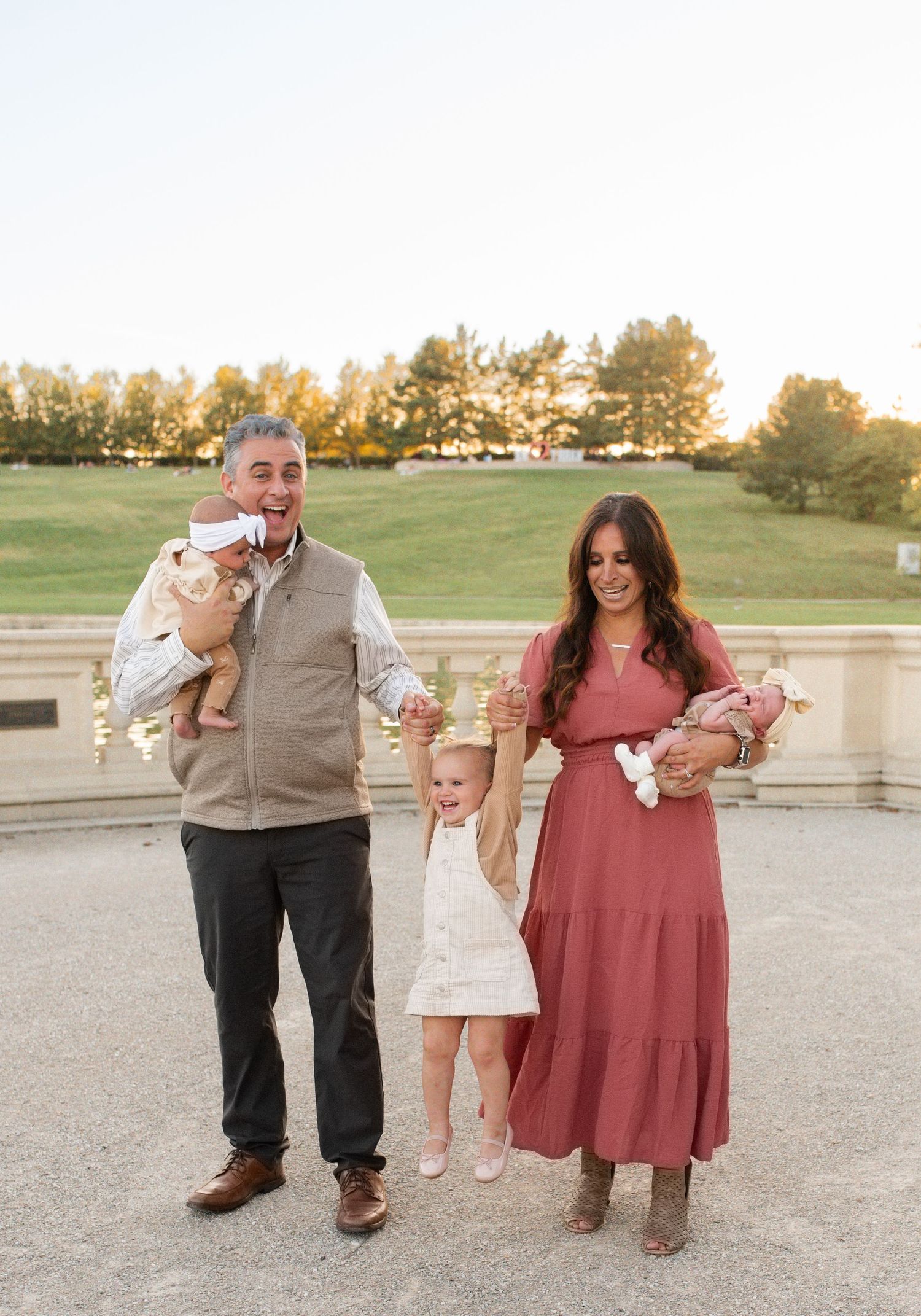 Family of four posing outdoors at golden hour, parents holding infants while toddler stands between them smiling.
