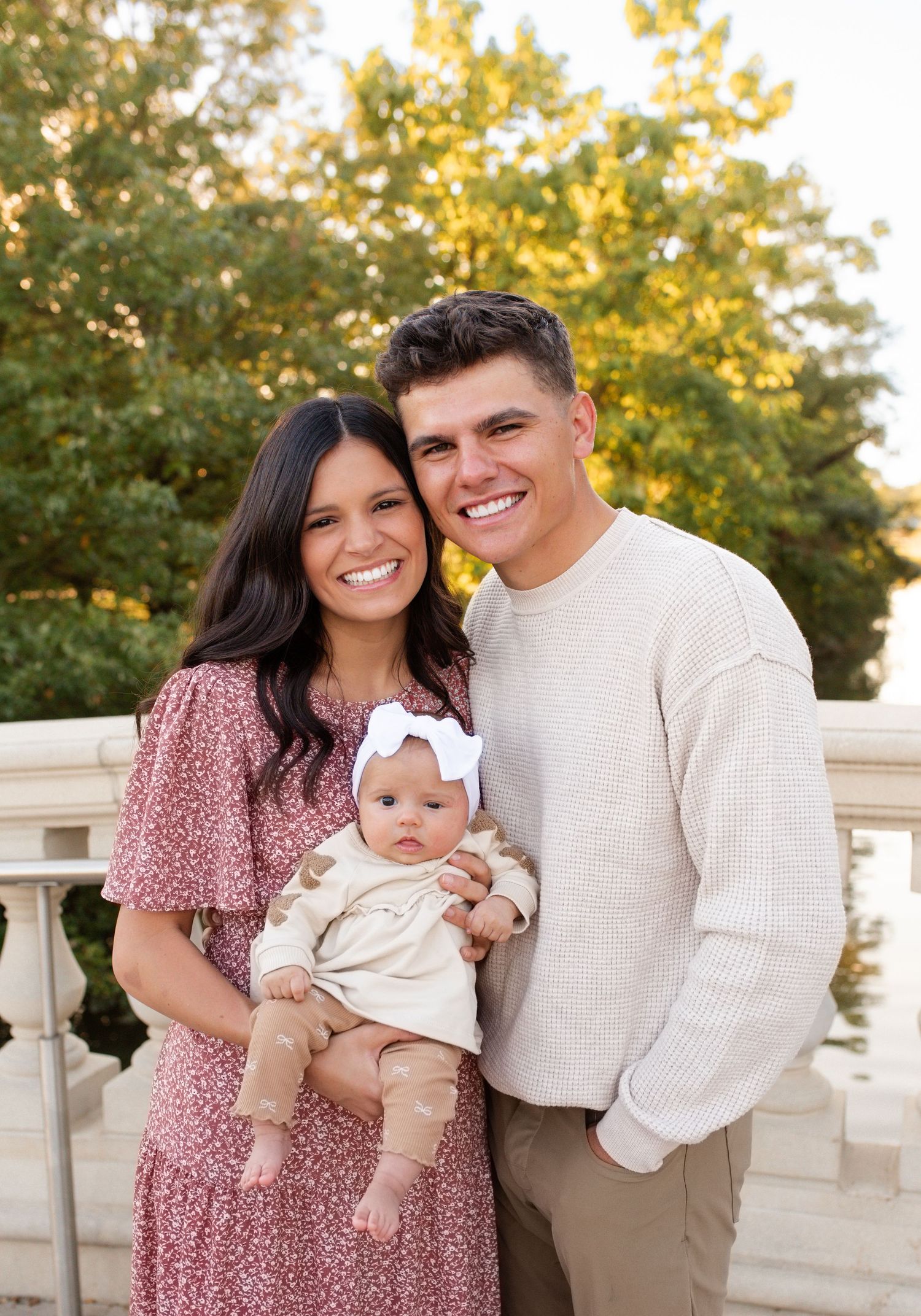 Young couple smiling outdoors holding their baby girl in a white bow, posing near stone railing with green trees behind in St. Louis