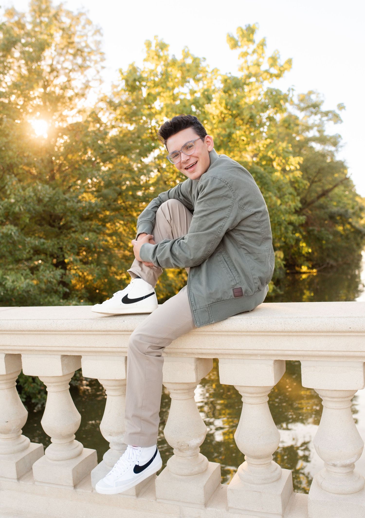 Young man in green jacket and Nike sneakers sits smiling on stone balustrade with sunlit trees behind him at Forest park in St. Louis