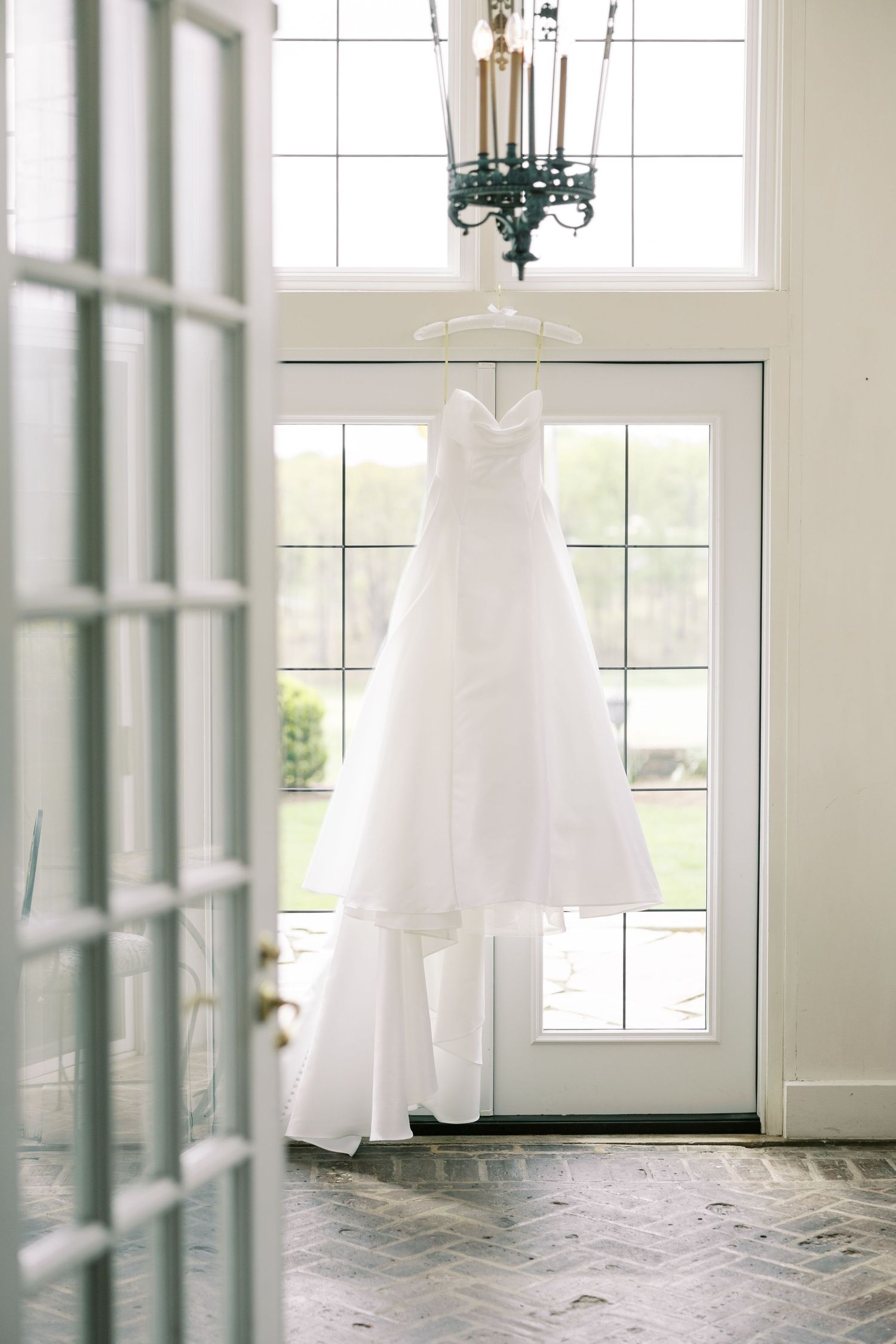 White strapless wedding dress hanging on a chandelier in a bright, elegant entryway with large windows.