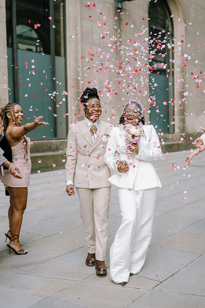 Two brides in white and cream suits walk through colorful heart confetti, celebrating their wedding day outdoors.