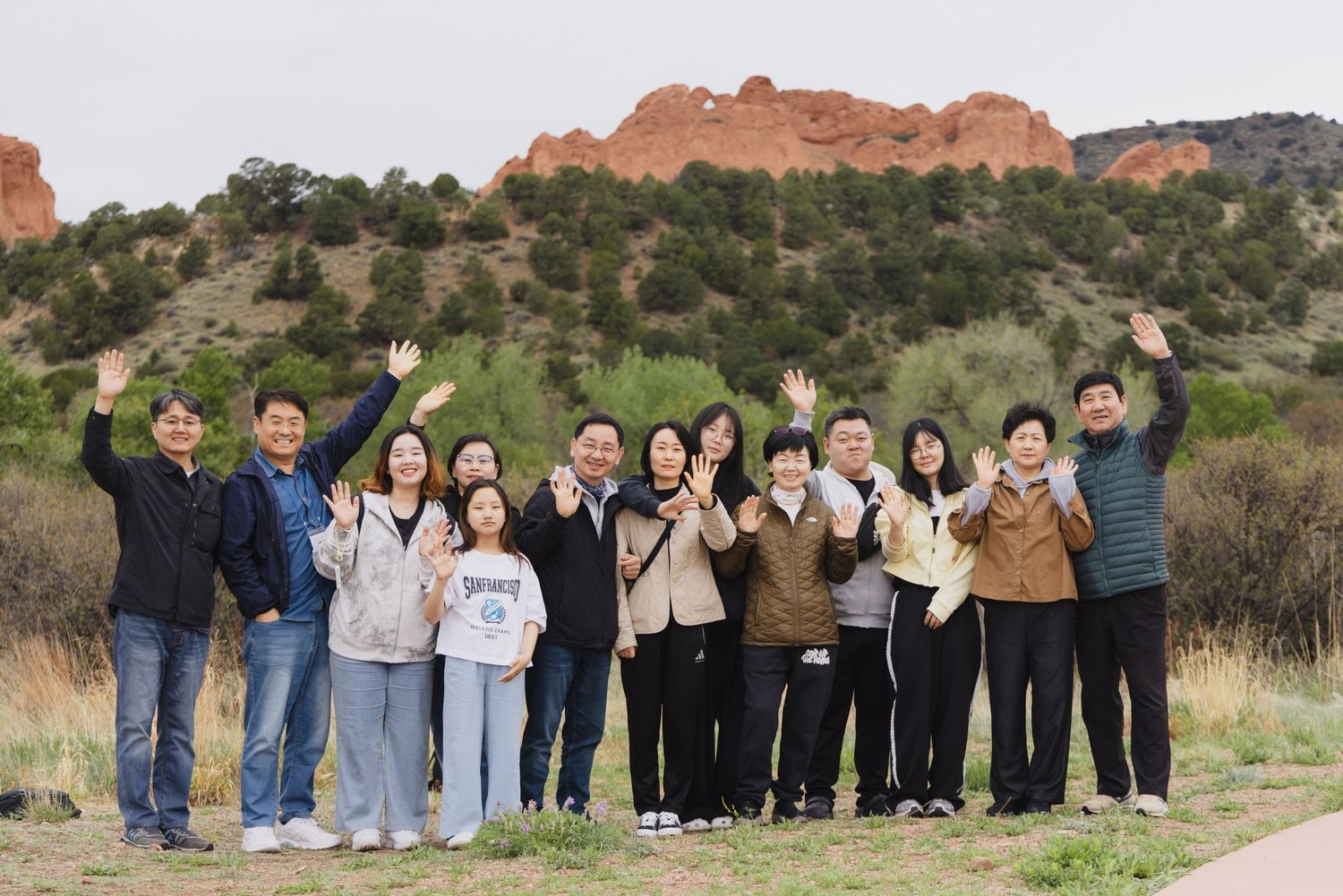 A large group of people waving and smiling outdoors with red rock formations and green hills in the background.
