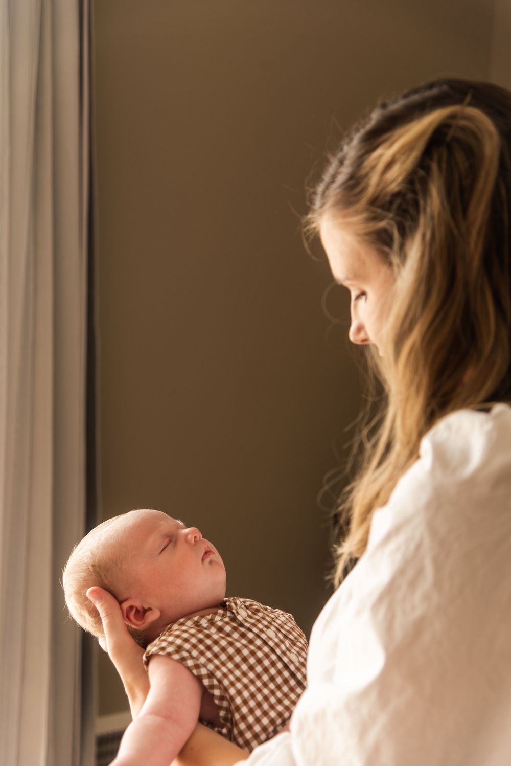 A young mother tenderly gazes at her newborn baby dressed in a brown checkered outfit near a sunlit window.