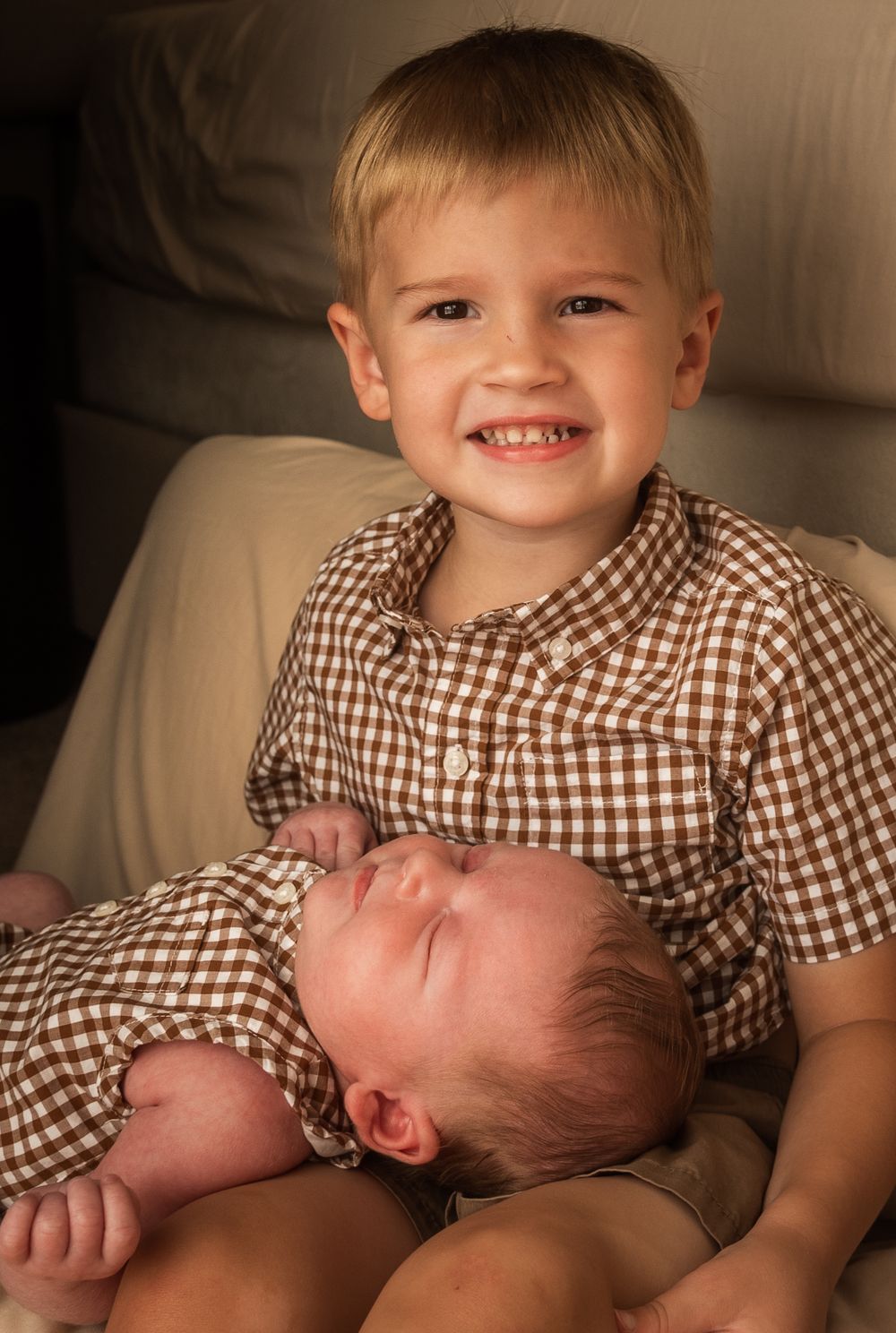 Smiling toddler boy in brown gingham shirt holds sleeping newborn baby sibling on a couch.