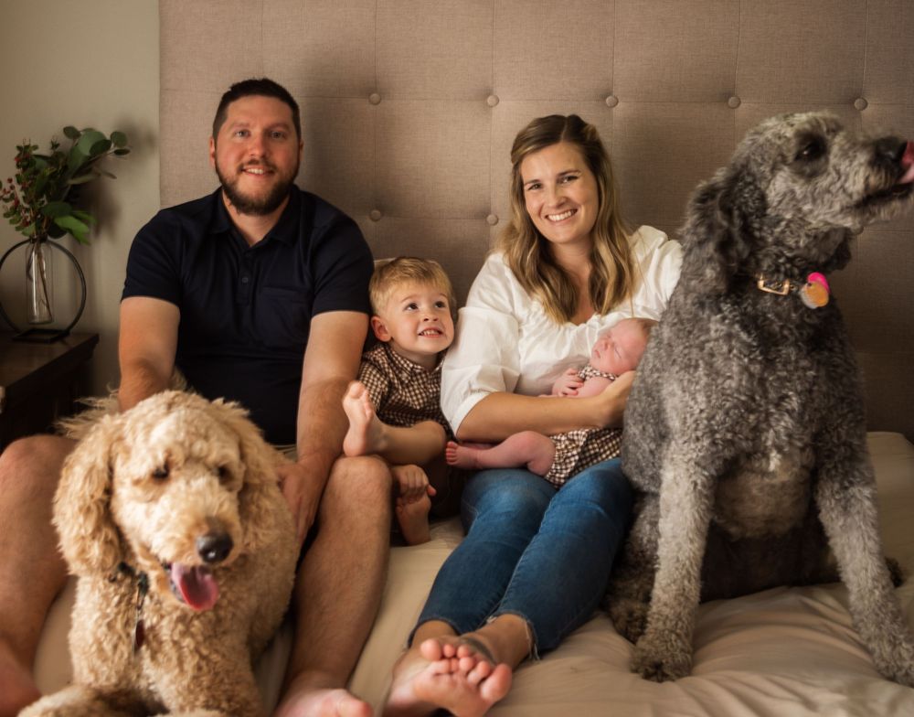 Happy family with two dogs, a toddler, and newborn baby sitting together on a bed in a warm home setting.