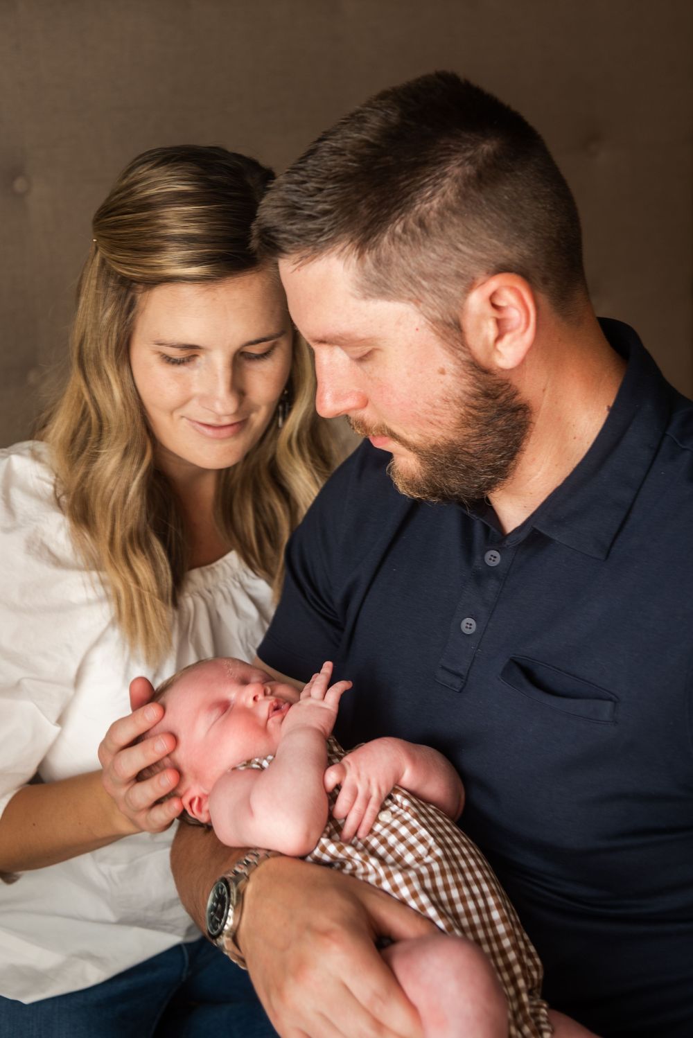 A couple tenderly gazes at their newborn baby, with the mother in white and father in navy holding the infant.