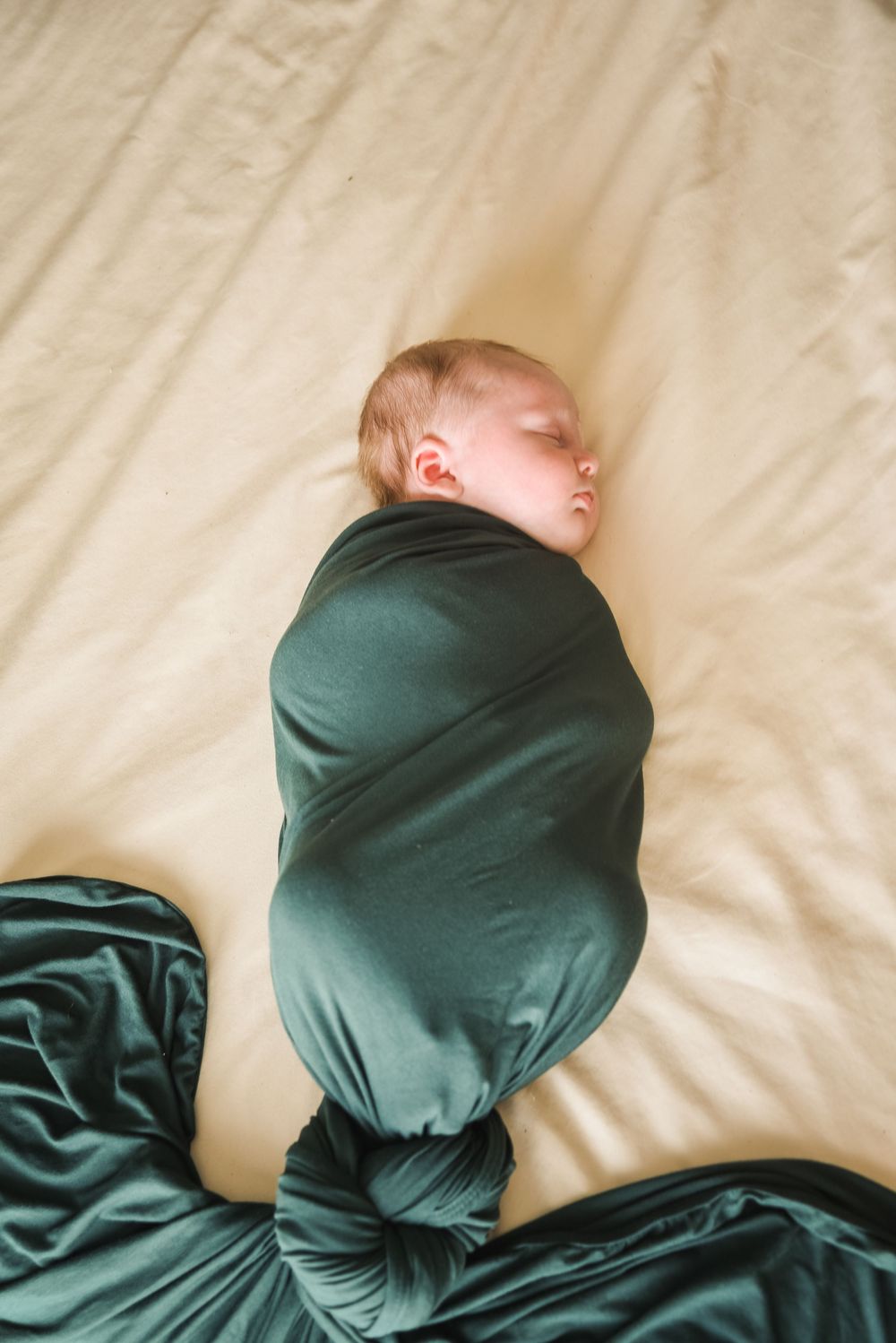 Newborn baby swaddled in a dark green blanket sleeping peacefully on a cream colored bed.