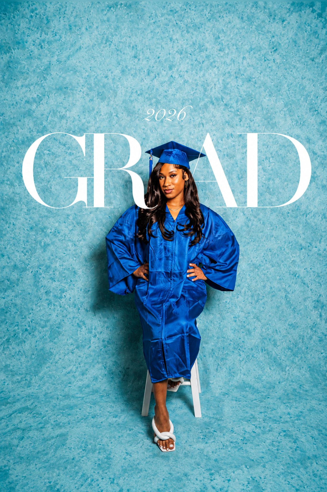 A young woman in a royal blue graduation cap and gown poses confidently on a stool against a blue backdrop with 2026 GRAD text.