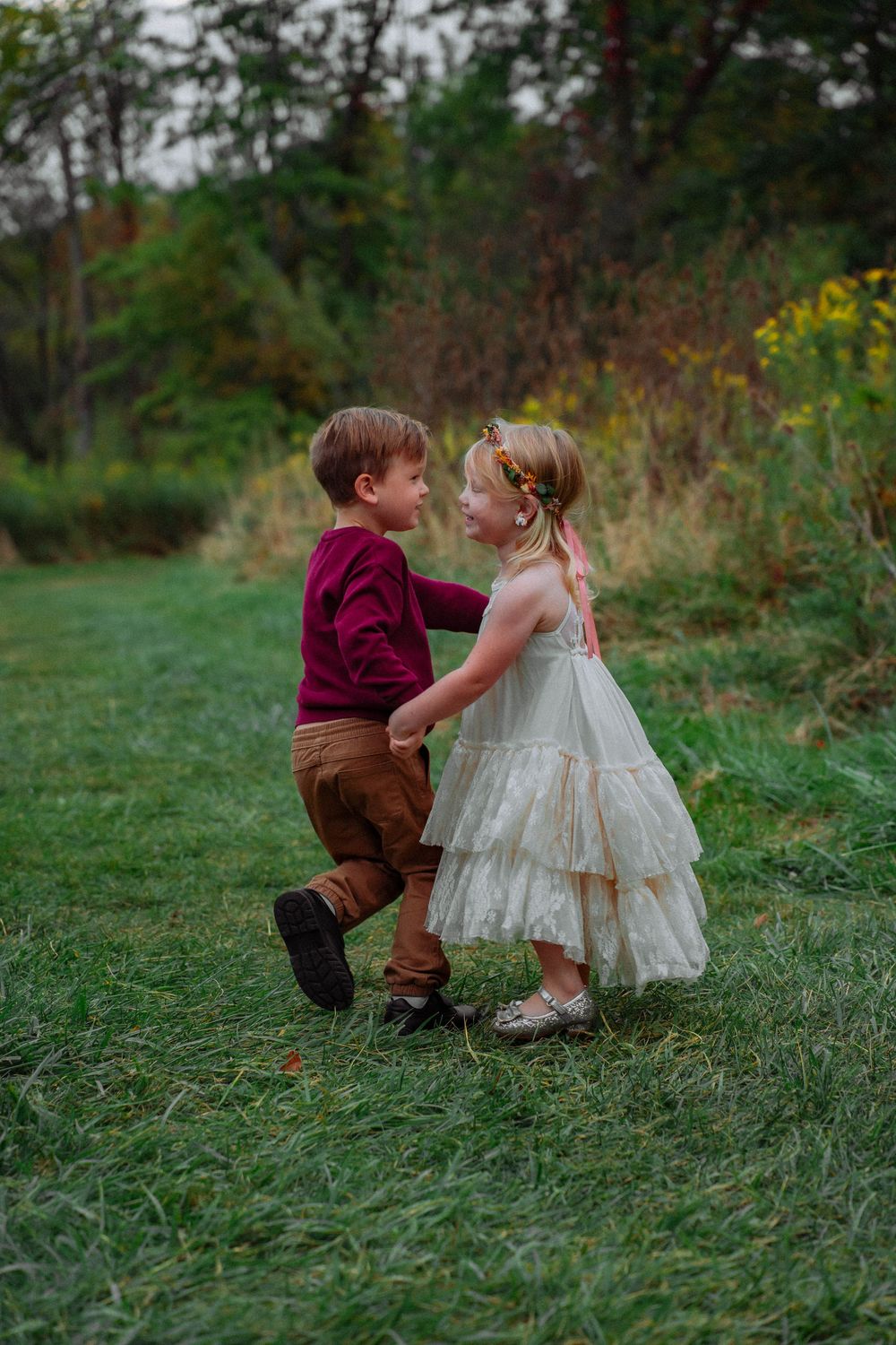 Two young children hold hands and dance together on a green grass path outdoors.