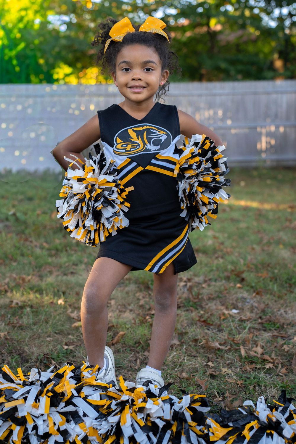 Young girl in black and gold cheerleader uniform poses with pom-poms outdoors.