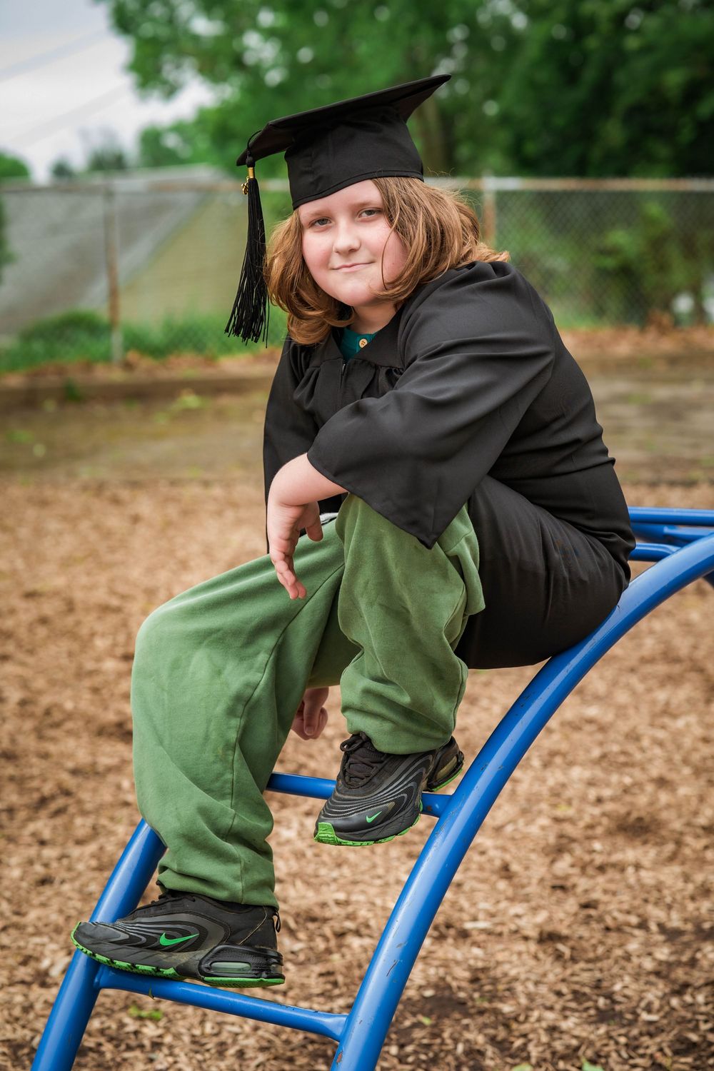 Child in graduation cap and gown sits on playground equipment smiling at camera.