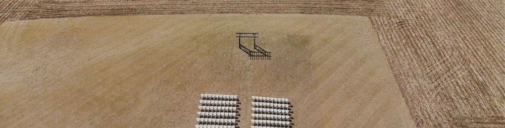 Aerial view of a large flat agricultural field with solar panels and a metal structure installed in the dirt.