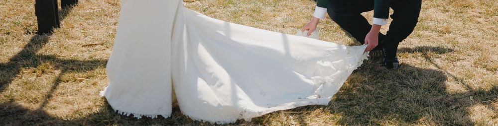 A person adjusts the train of a white wedding dress on a bride standing on green grass outdoors.