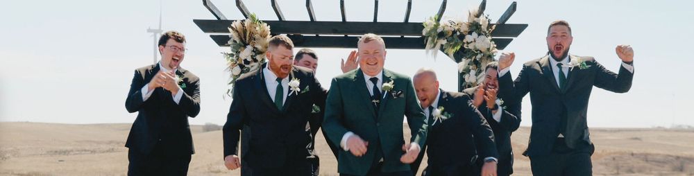 Groomsmen in suits celebrate outdoors at a beach wedding ceremony with a floral arch backdrop.