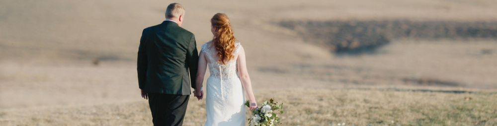 A bride and groom walk hand in hand through a sunlit open field, the bride wearing a flowing white gown holding a bouquet.