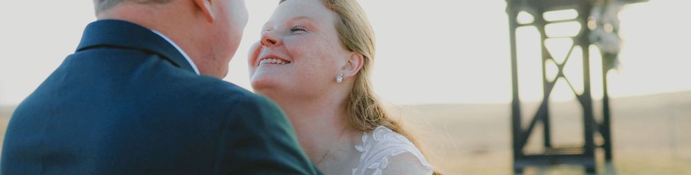 A bride smiles joyfully at her groom during an outdoor wedding ceremony at golden hour.