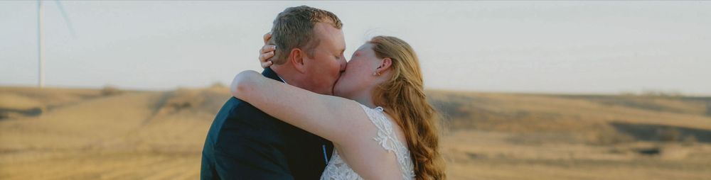 A couple shares a romantic kiss at golden hour in an open wheat field with a windmill in the background.