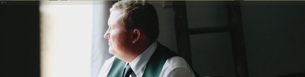 A groom in a gray vest and tie gazes thoughtfully out a window in soft natural light.