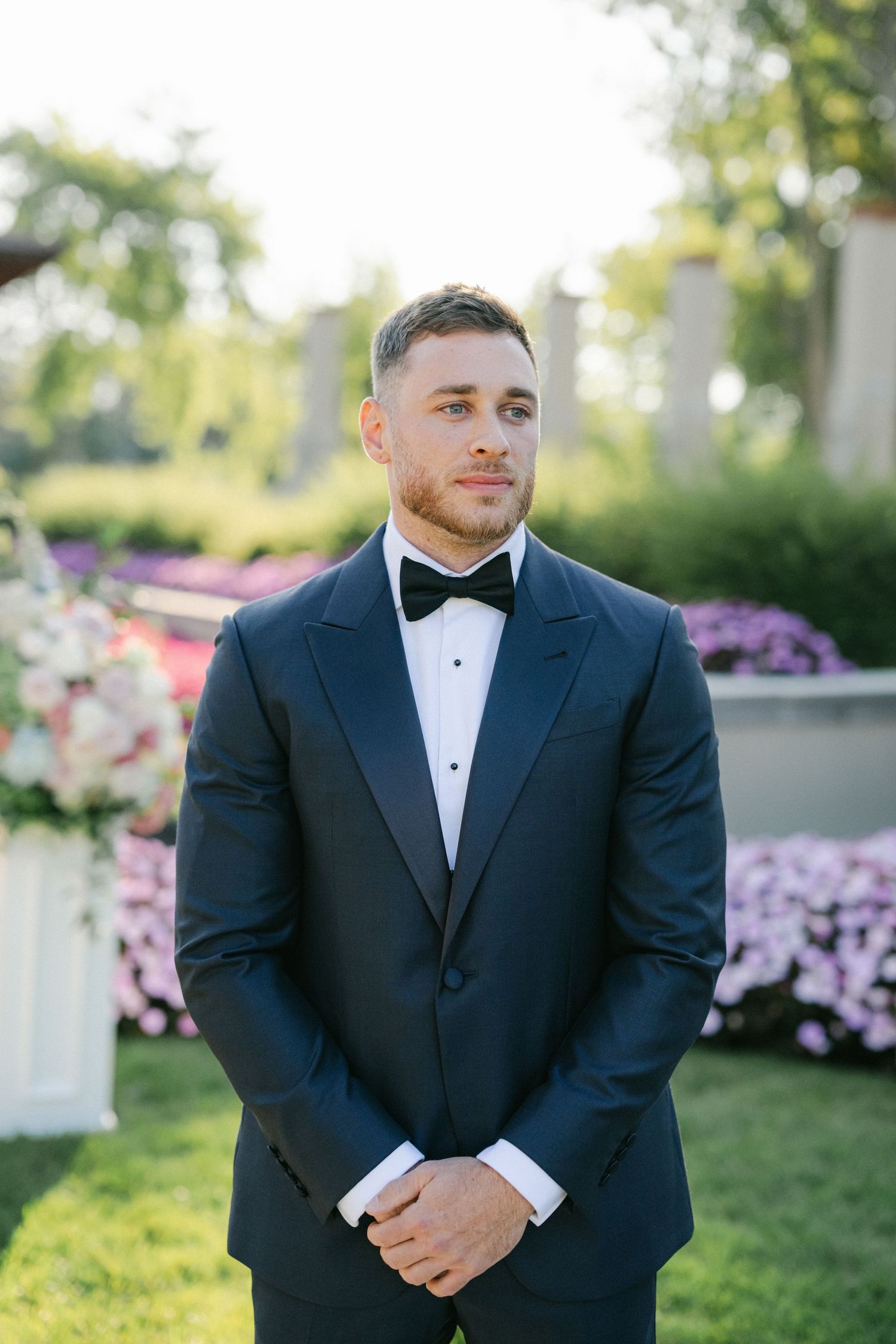 Armour House Wedding - A groom in a navy tuxedo with black bow tie stands outdoors at a garden wedding ceremony.