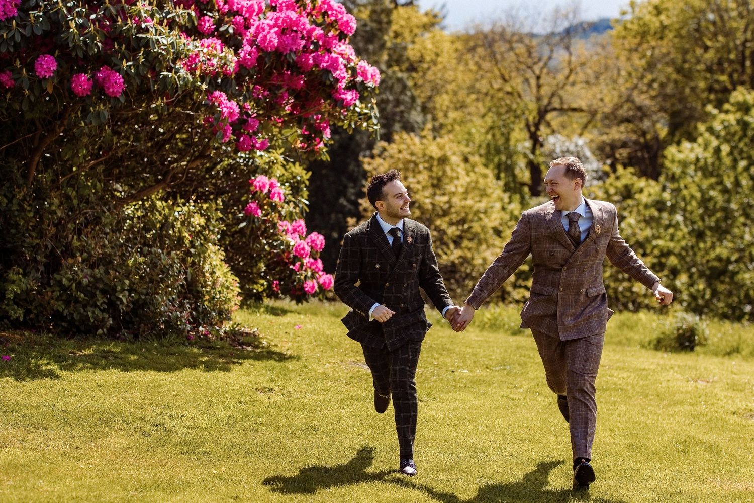 Two grooms in suits hold hands running through a sunny garden with vibrant pink rhododendrons blooming nearby.