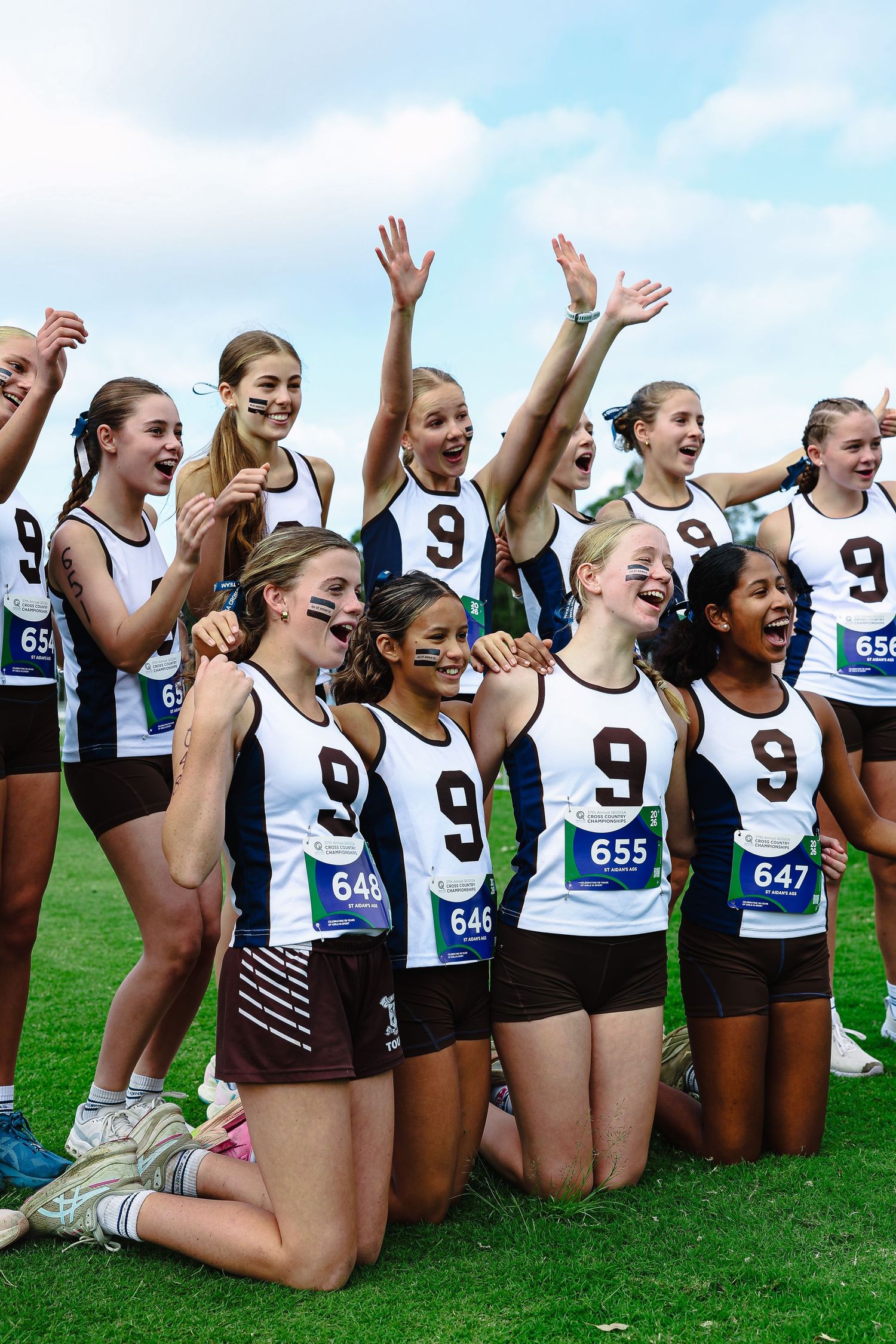 Young female athletes in blue uniforms race at the start of a track meet on a green grass field.